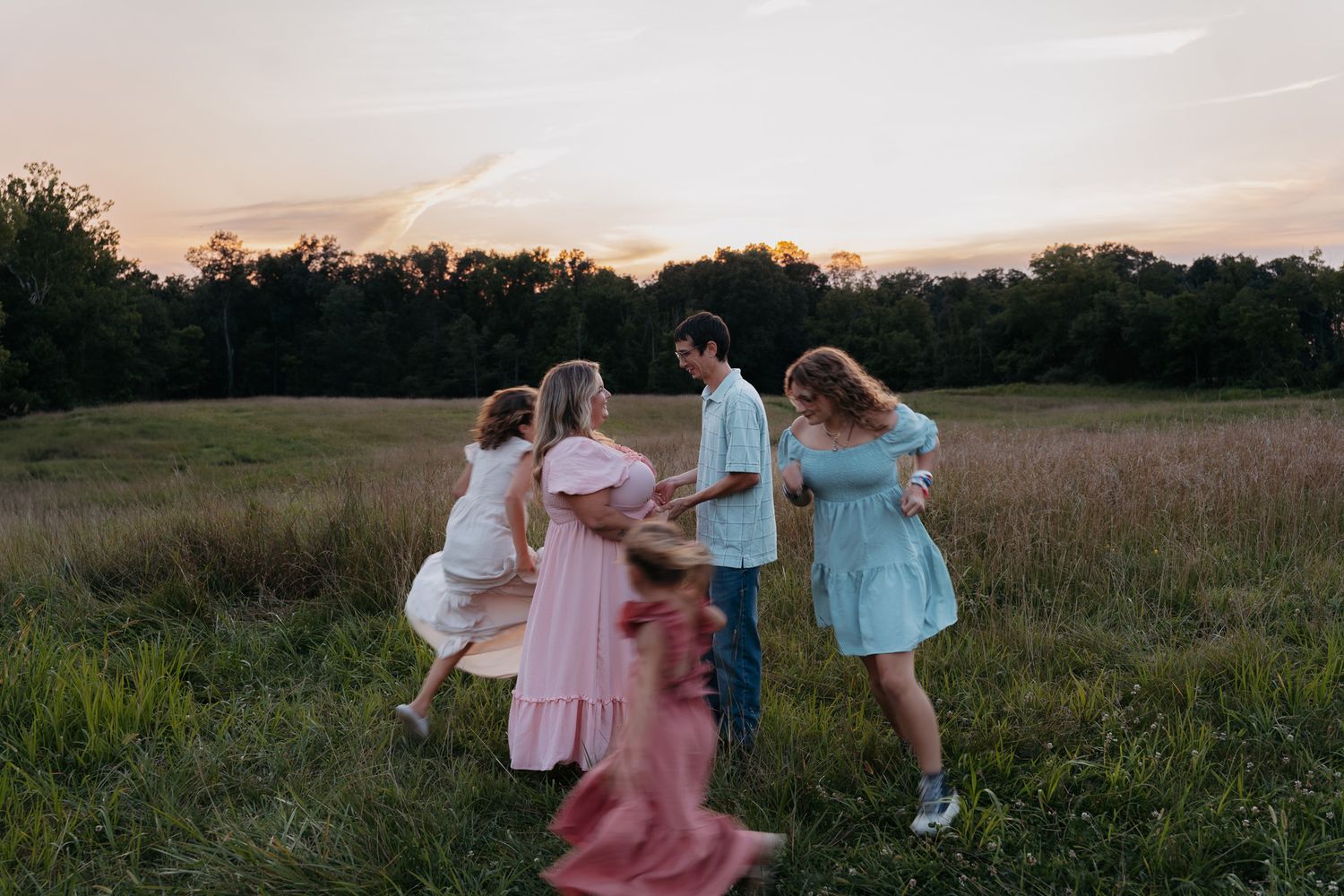 Family dancing together in a golden field at sunset, wearing pastel dresses and casual attire.