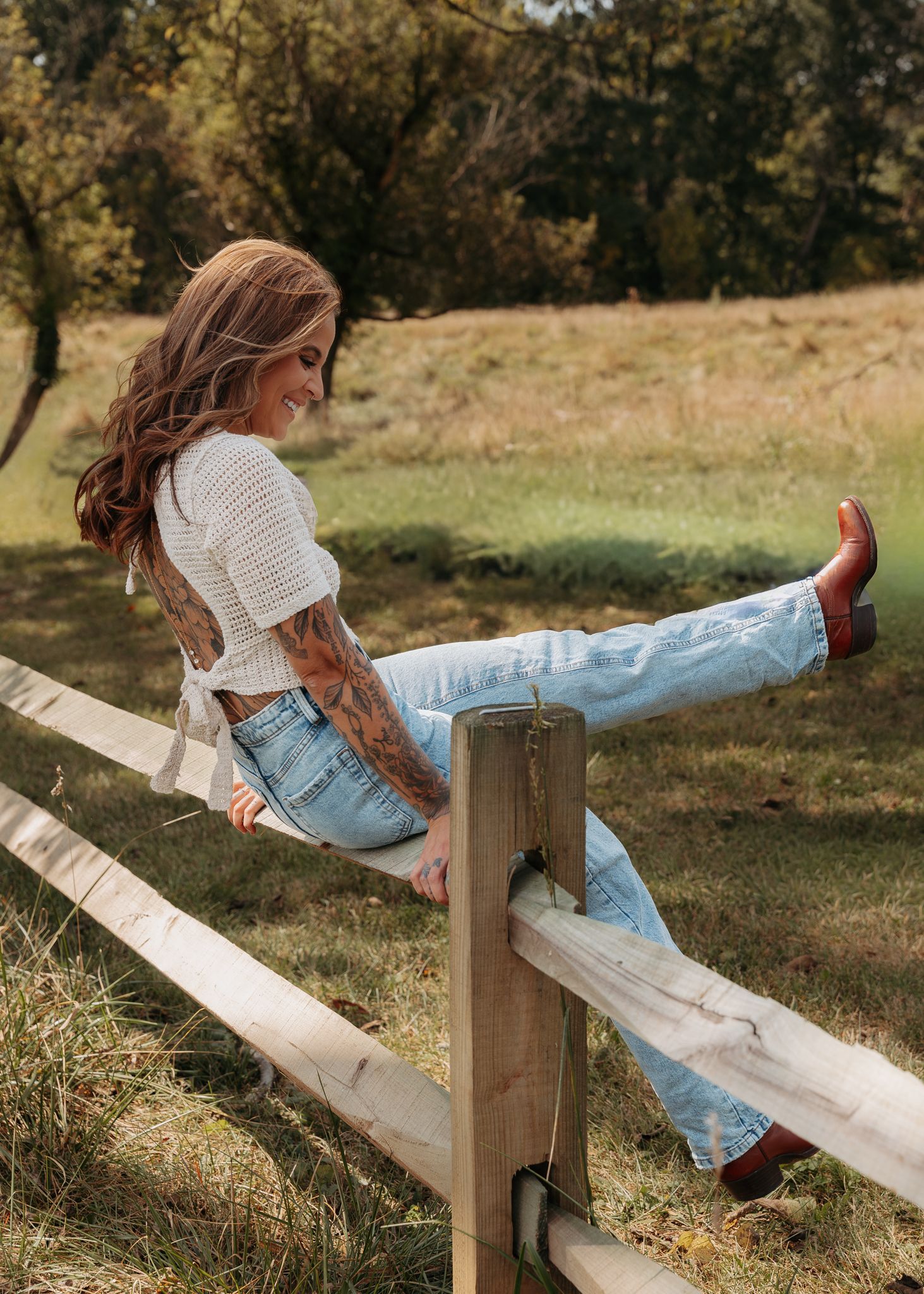 Young woman with red hair sitting on a wooden fence in jeans and cowboy boots in a field.
