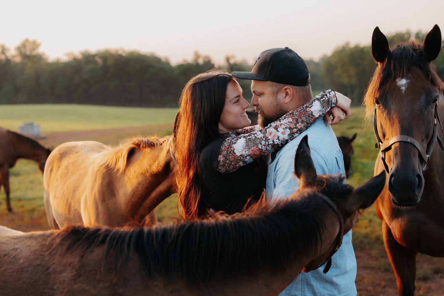 Romantic couple sharing a tender moment surrounded by horses in a golden field at sunset.