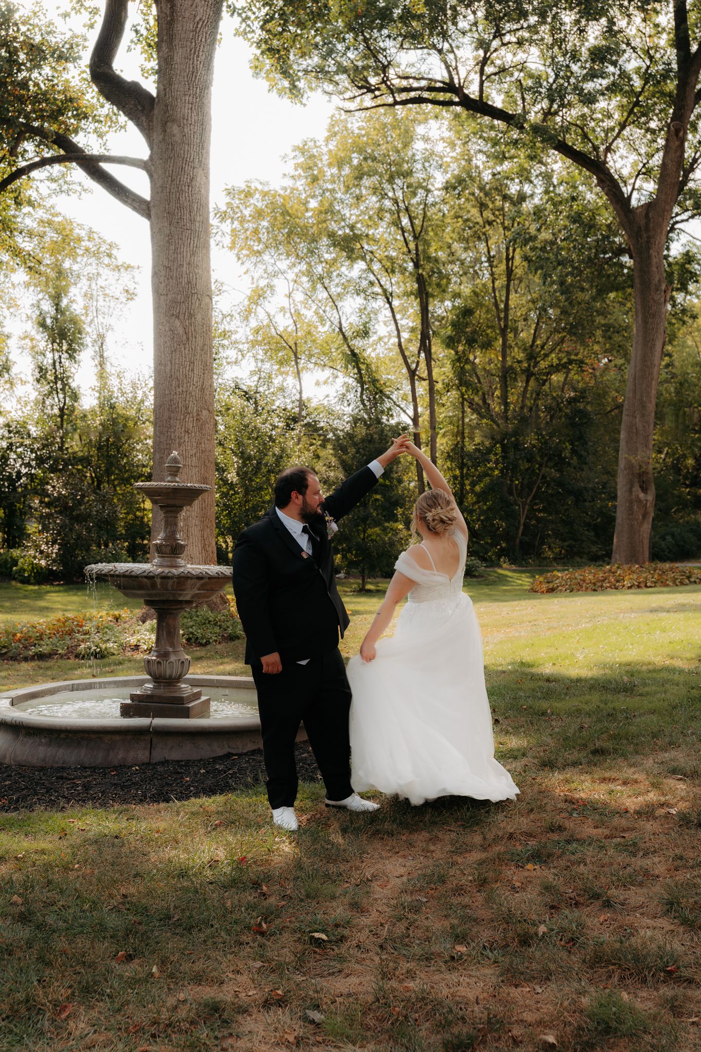 Groom twirling bride in white off-shoulder gown near stone fountain in sunlit outdoor garden setting.