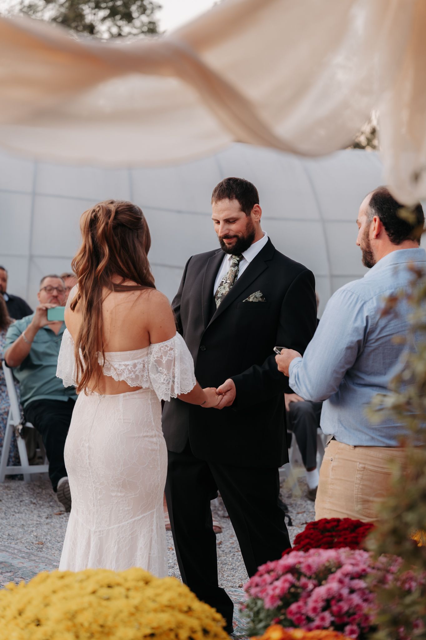 Groom in dark suit holding bride's hands during outdoor wedding ceremony with flowers.