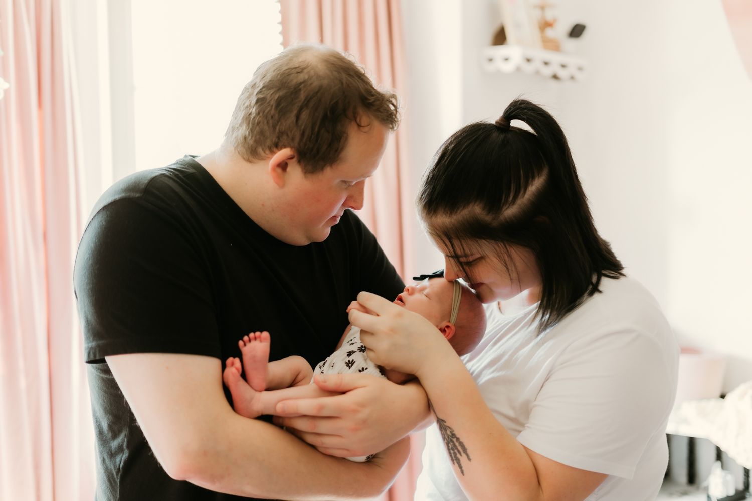 New parents tenderly hold their newborn baby in a softly lit pink nursery room together.