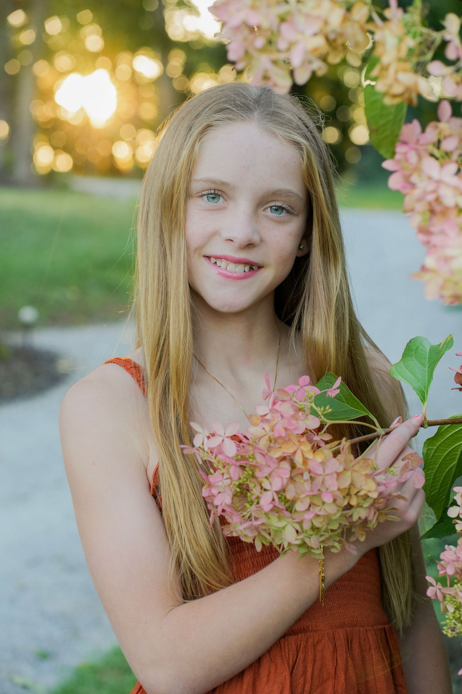 Smiling blonde girl in orange dress holds pink hydrangea flowers in golden sunset light.