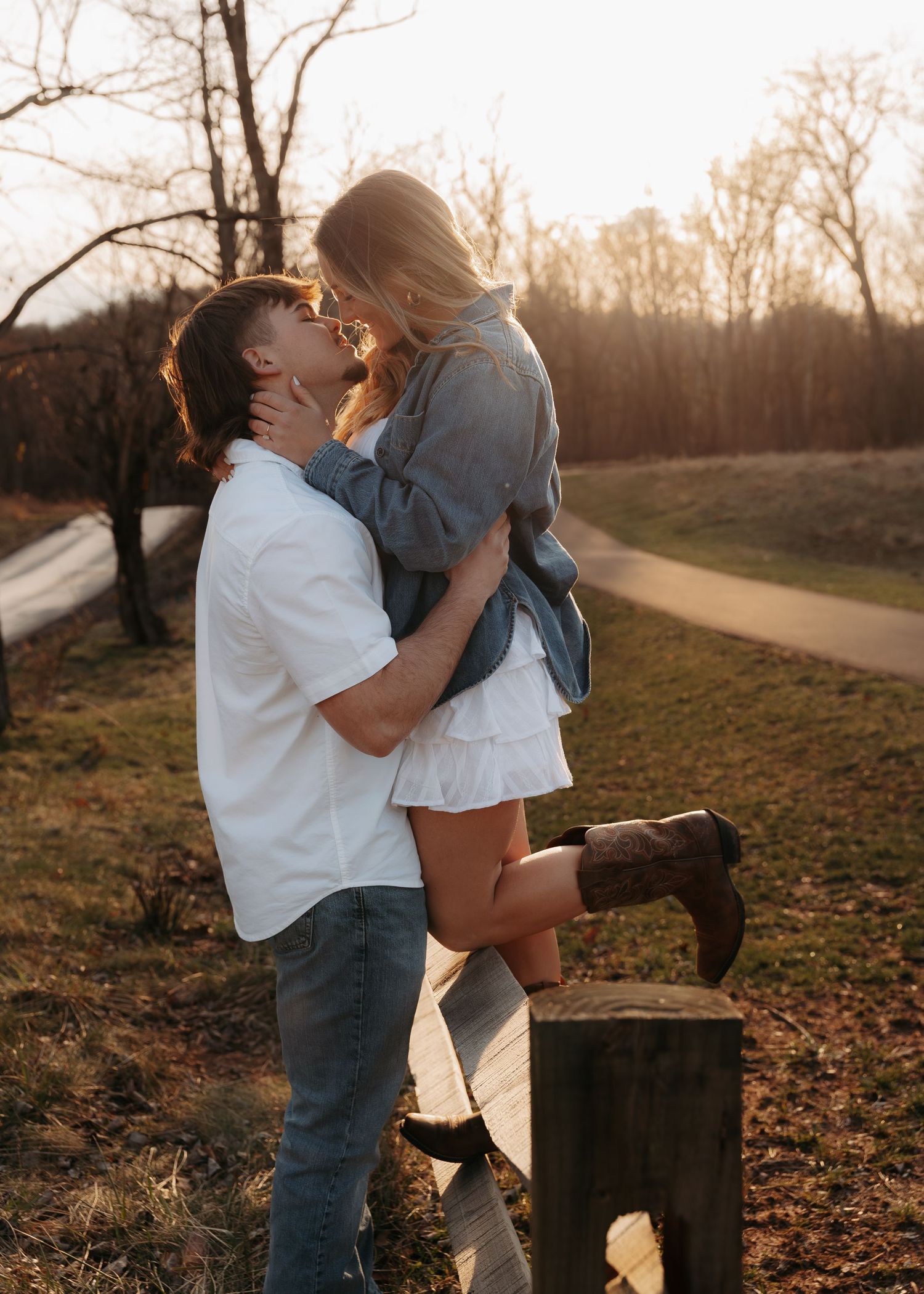 Couple sharing a romantic kiss at golden hour in a scenic outdoor setting with bare trees.