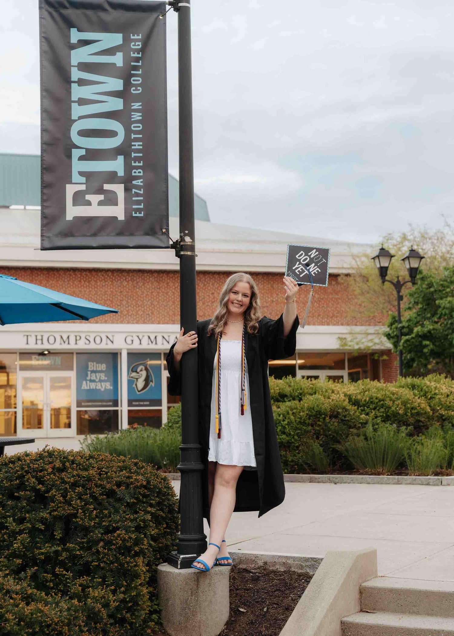 Female graduate in cap and gown poses by Elizabethtown College banner outside Thompson Gymnasium.