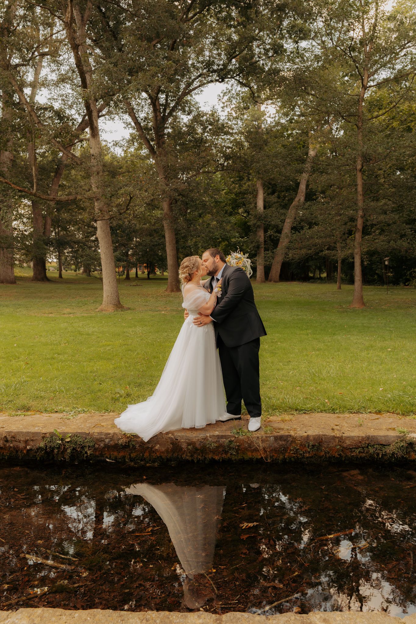 Bride and groom share a kiss on stone bridge reflected in still water surrounded by lush green trees.