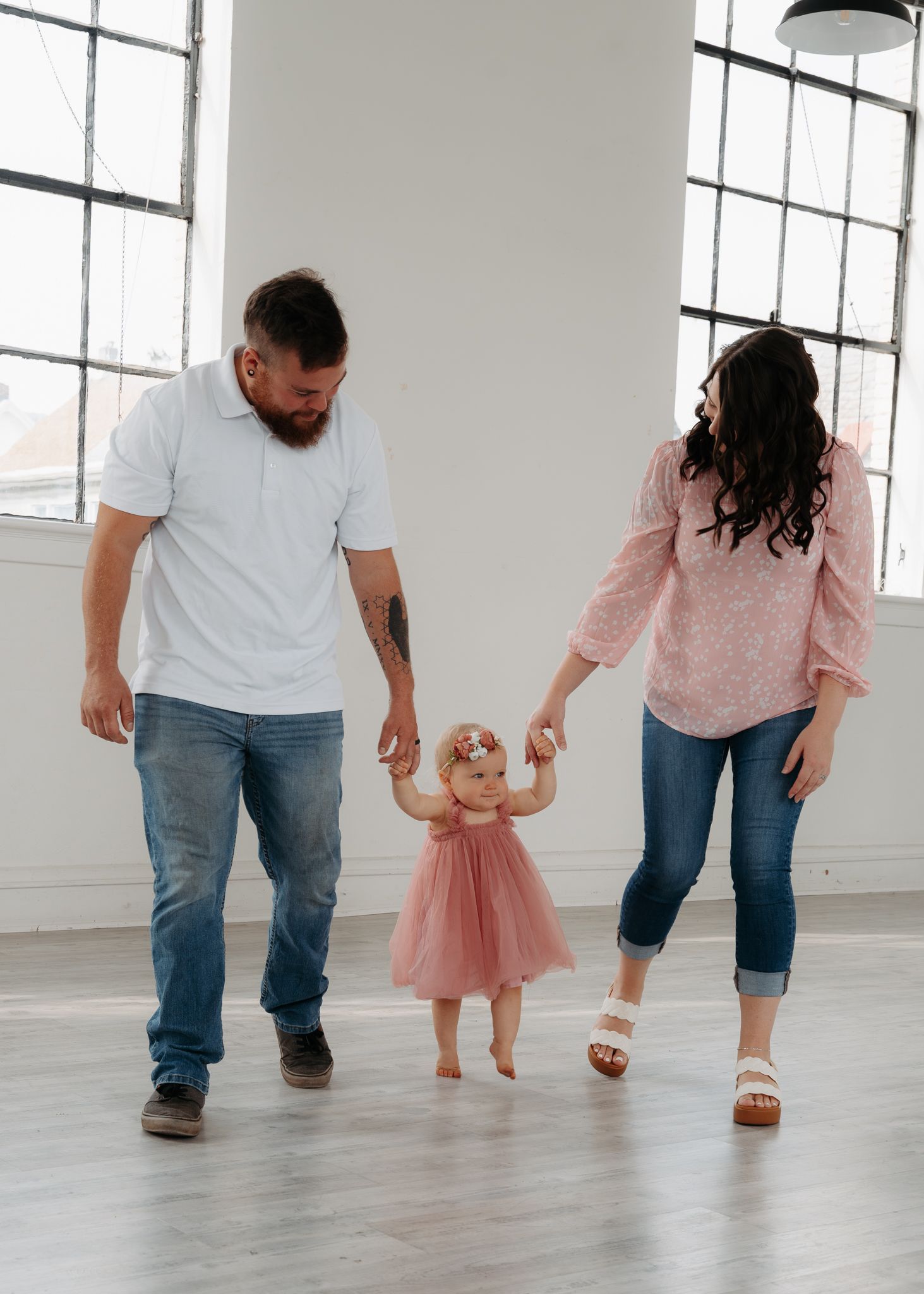 A young family holds hands with their toddler daughter in a bright white studio, all dressed in coordinating outfits.