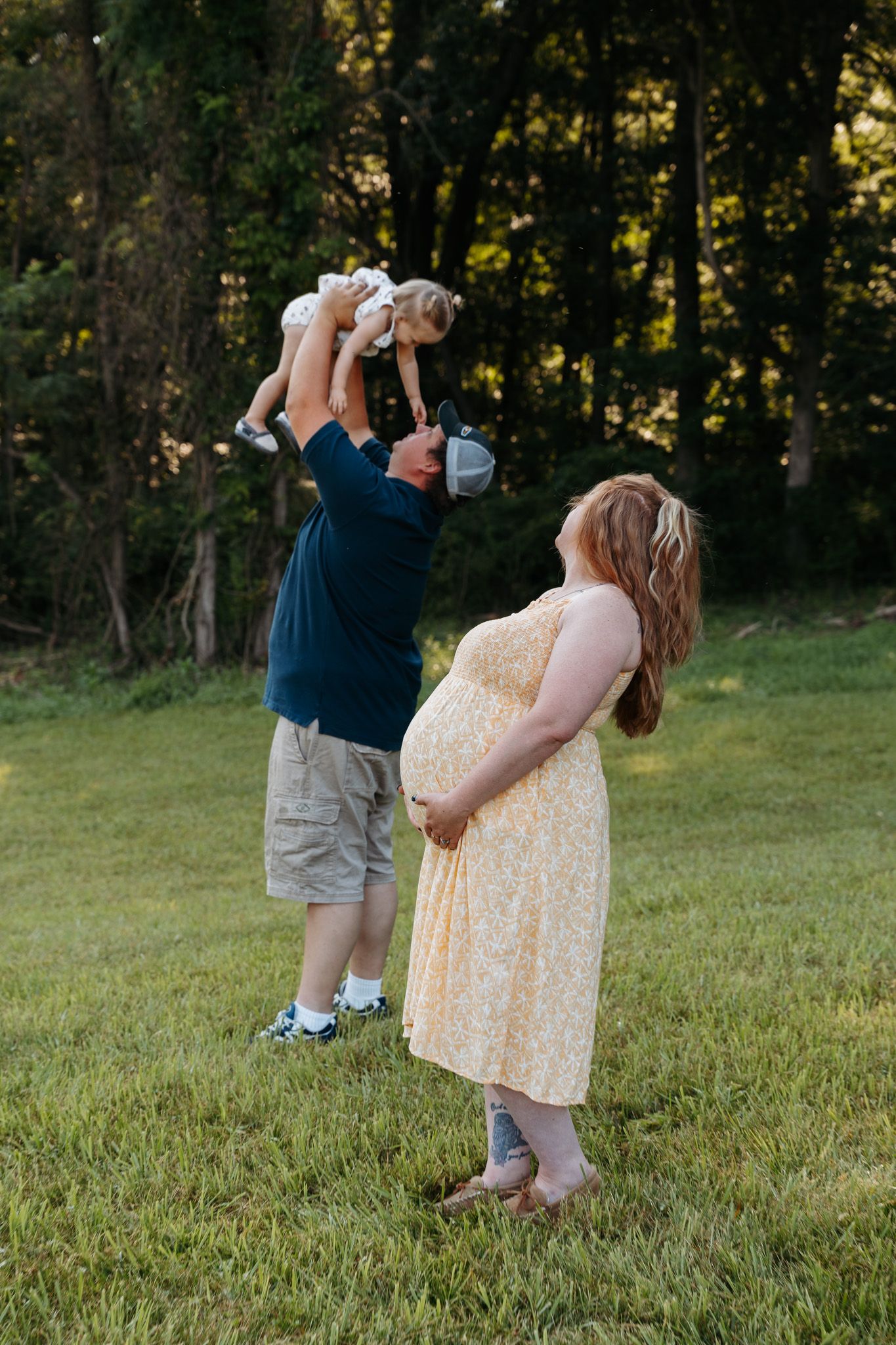 Pregnant woman in yellow dress watches partner lift toddler in sunny backyard.