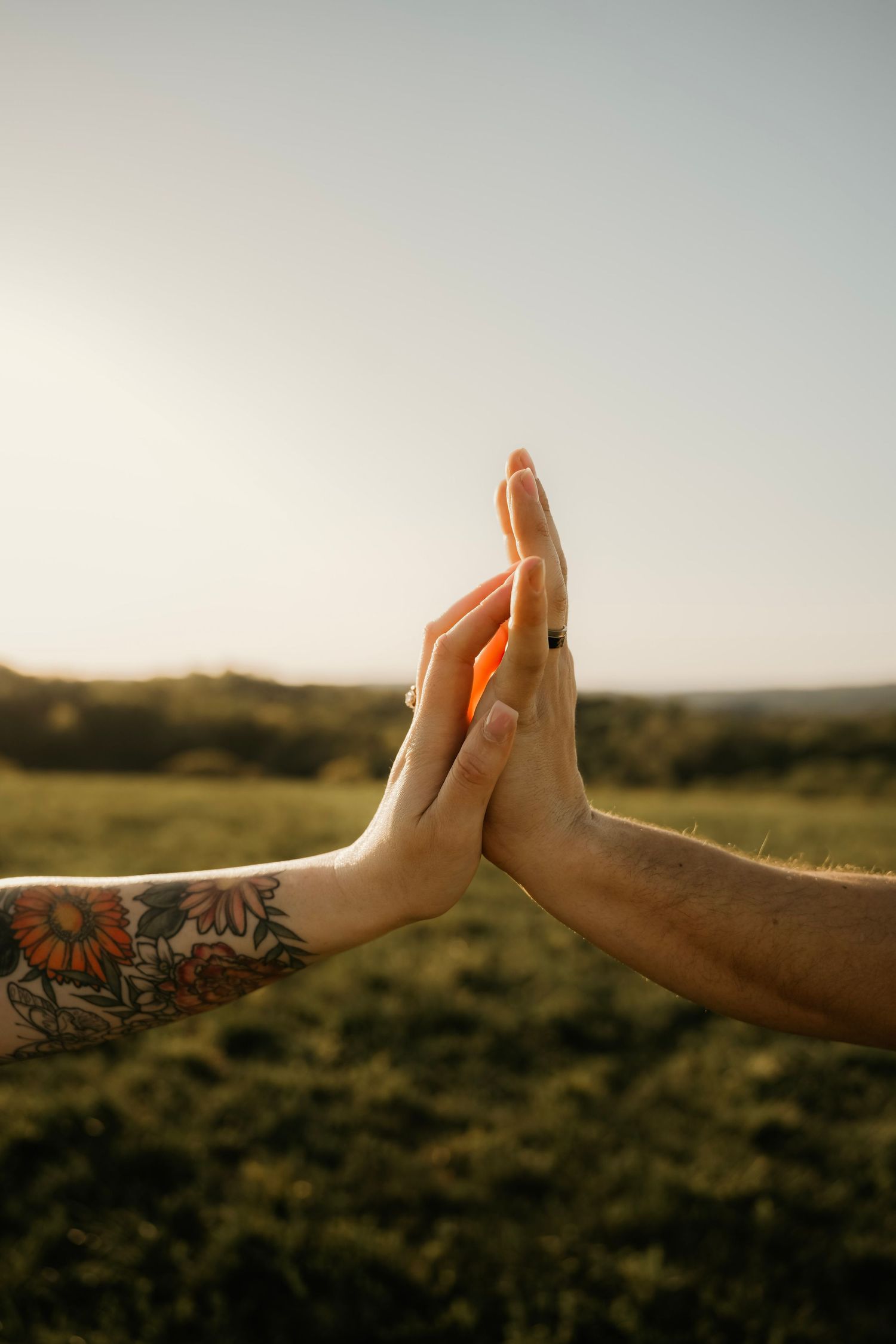 Two hands with tattoos pressing together against a golden sunset over a green field.