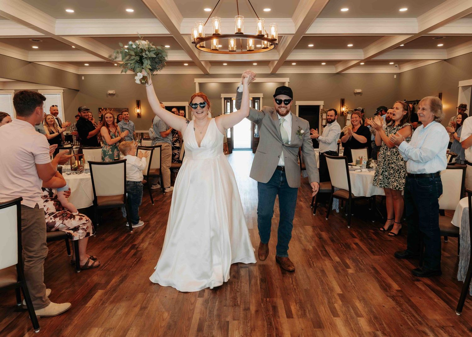 Bride and groom wearing sunglasses raise hands triumphantly while entering wedding reception to applauding guests.