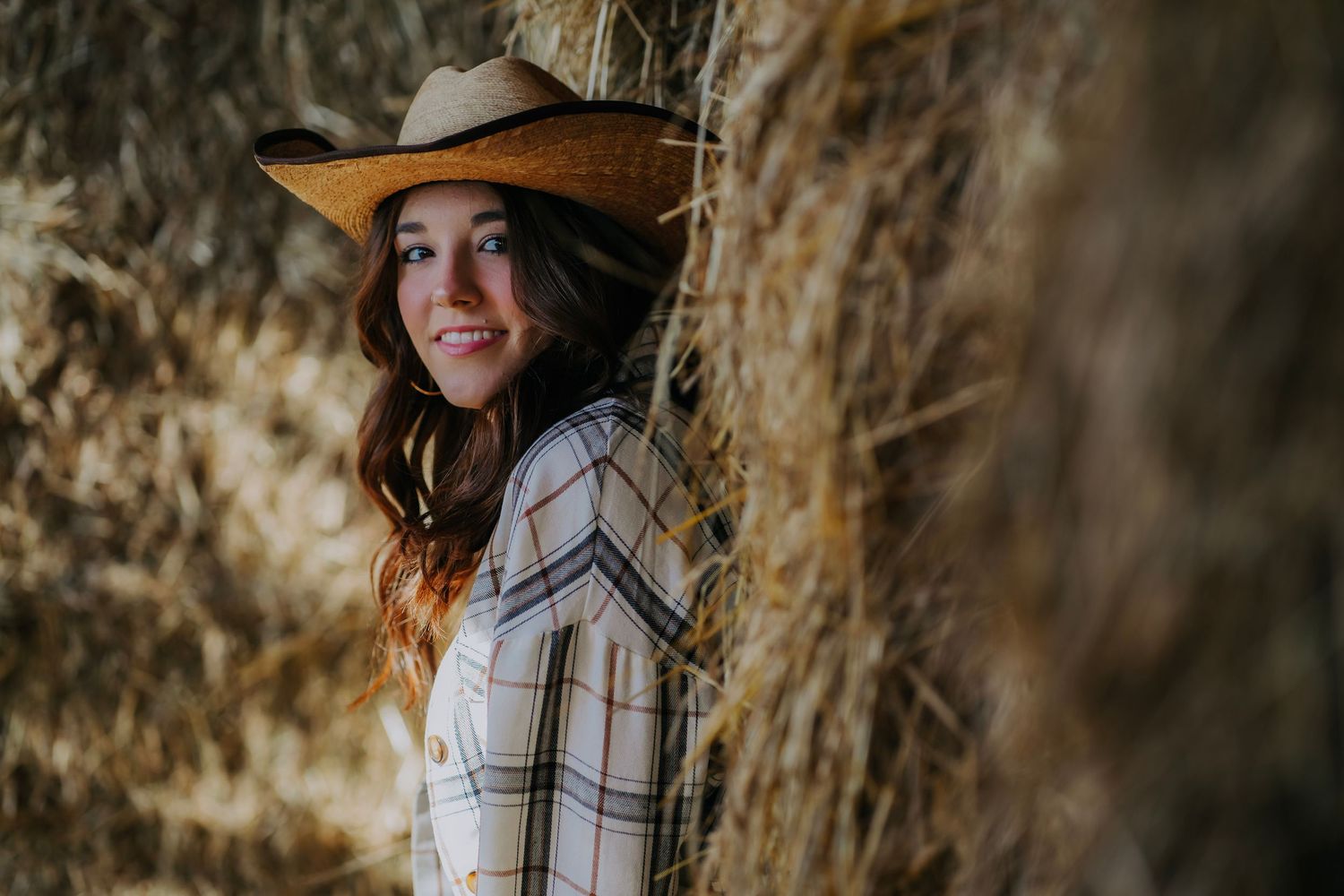 Young woman in cowboy hat smiling beside hay bales in rustic outdoor setting.