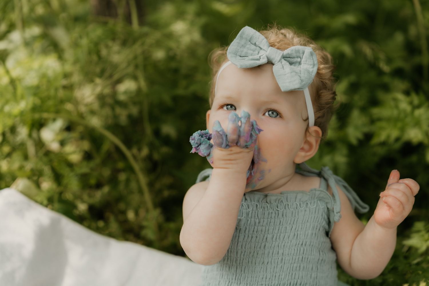 Cute toddler girl with a blue bow headband chews on a toy while sitting outdoors among green foliage.