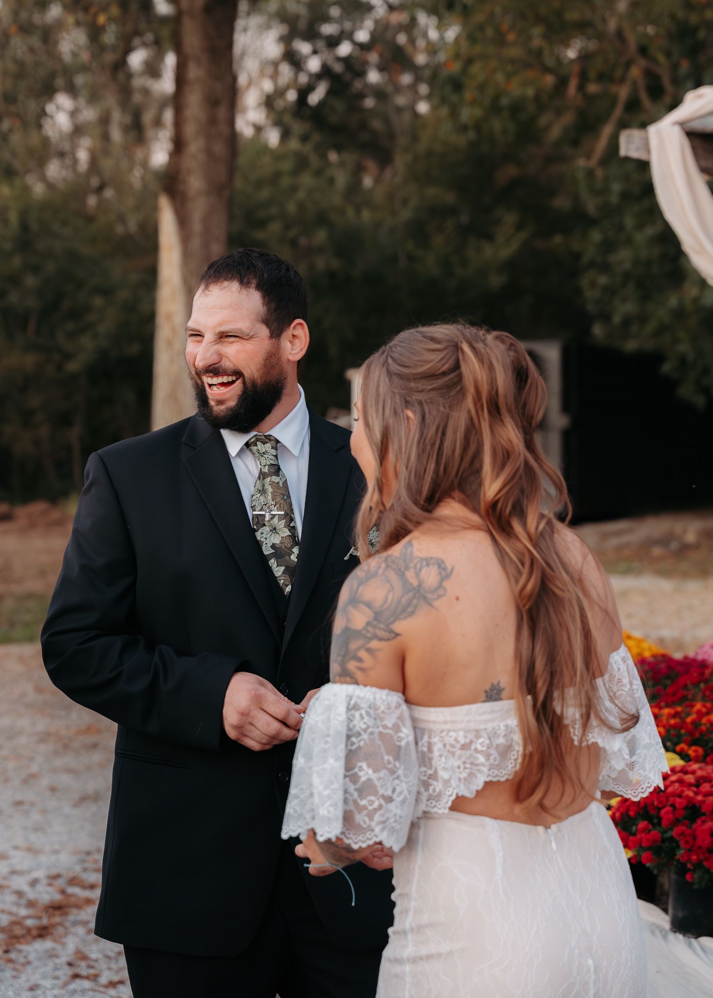 Groom in black suit laughing with tattooed bride in lace off-shoulder wedding dress outdoors.