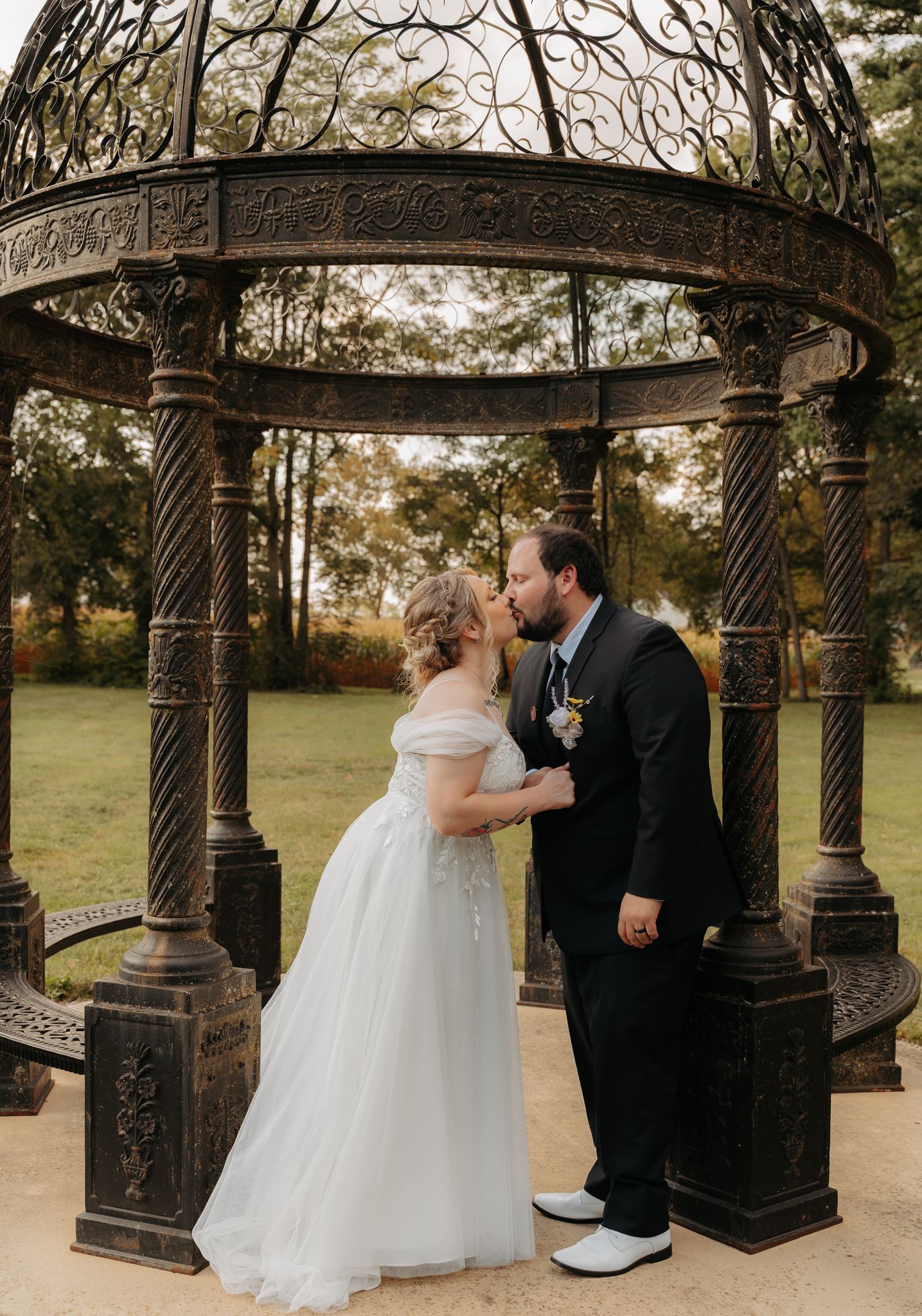 Bride and groom share a kiss under ornate iron gazebo with autumn foliage in background.