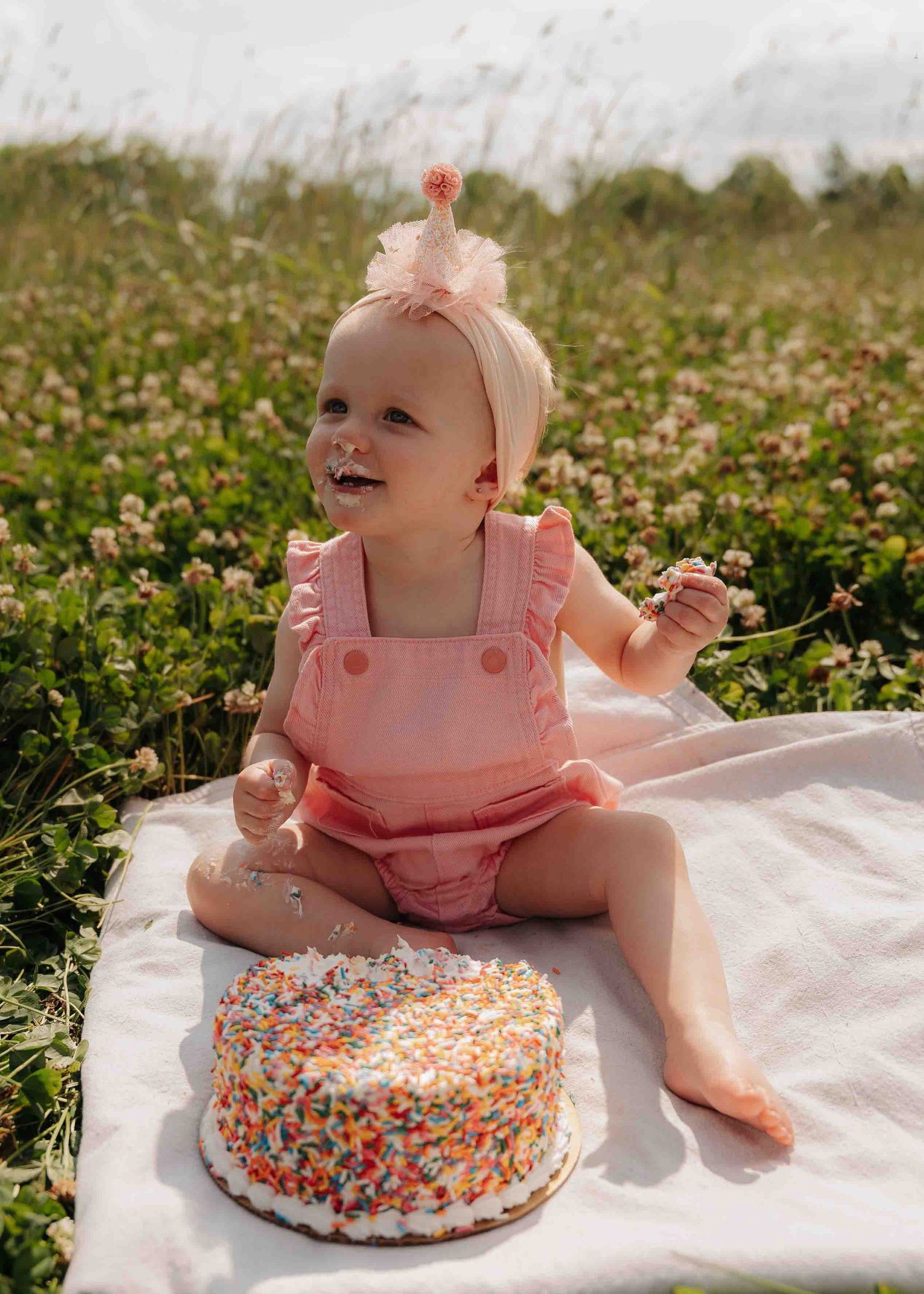 Happy baby in pink romper smashing a sprinkle cake in a wildflower field.