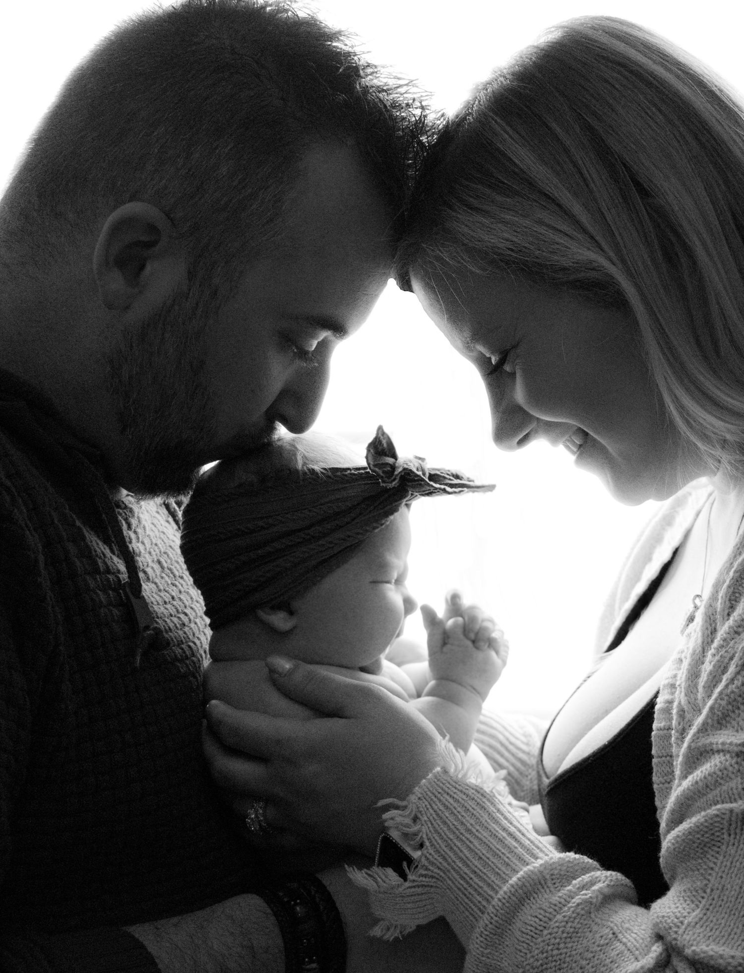 A black and white portrait of new parents lovingly gazing down at their sleeping newborn baby together.