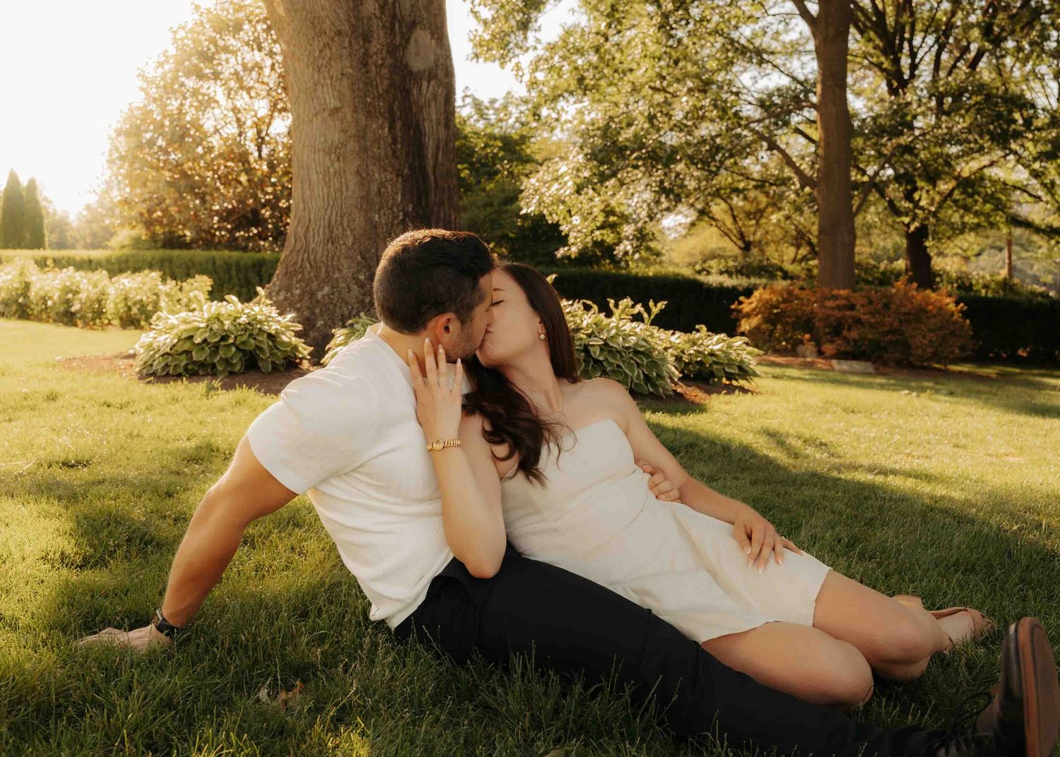 Couple kissing while relaxing on grass beneath a large tree in a beautiful sunlit garden.