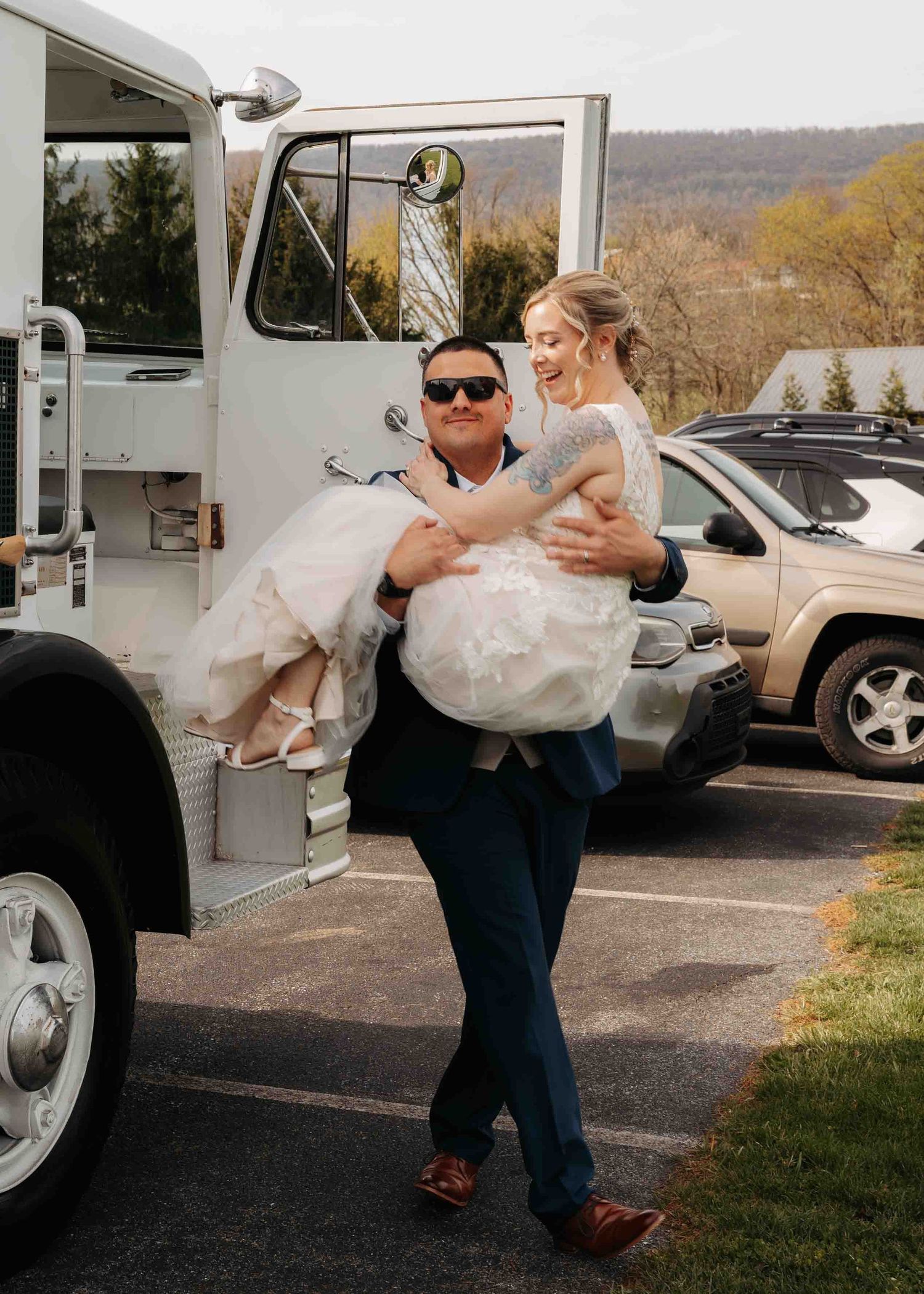 Groom in navy suit carries laughing bride in white gown beside vintage white fire truck at wedding.
