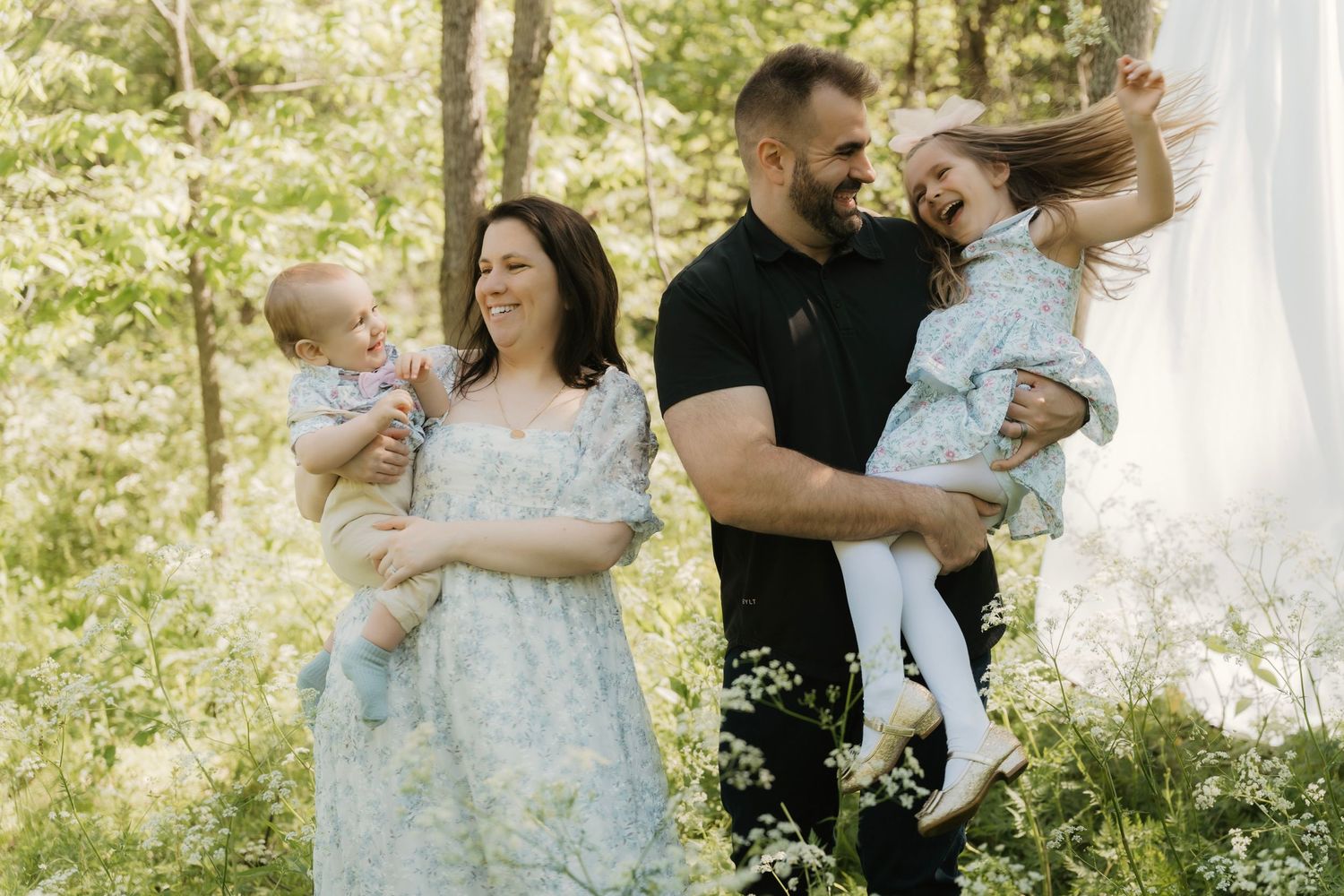 Happy family with two young children laughing outdoors among white wildflowers in bright forest.