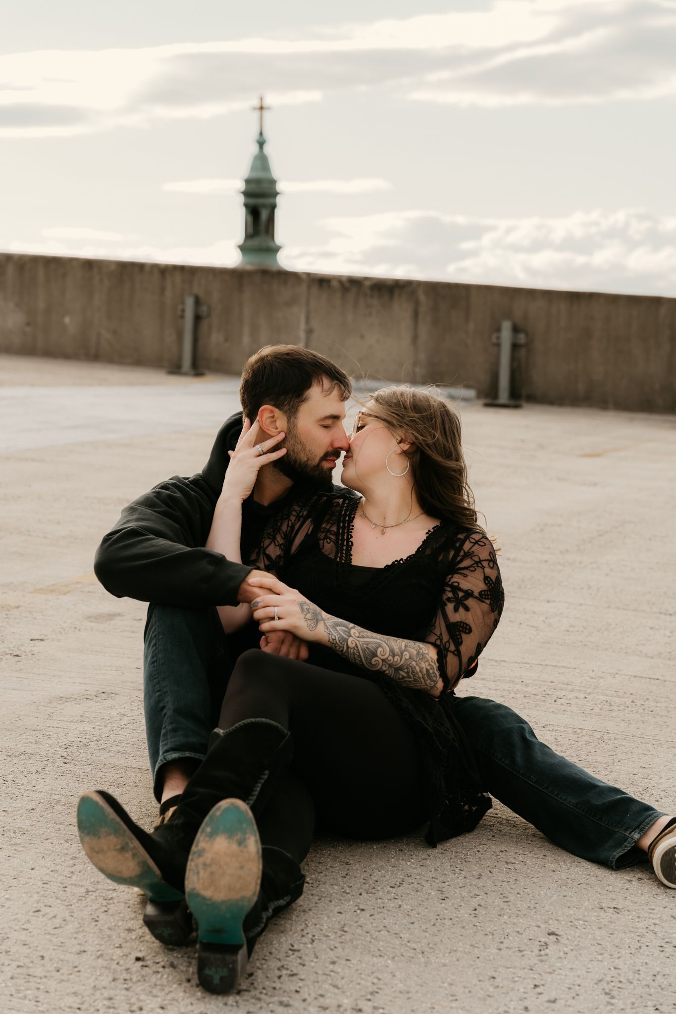 Tattooed couple sharing a kiss while sitting on a rooftop with a church steeple behind.