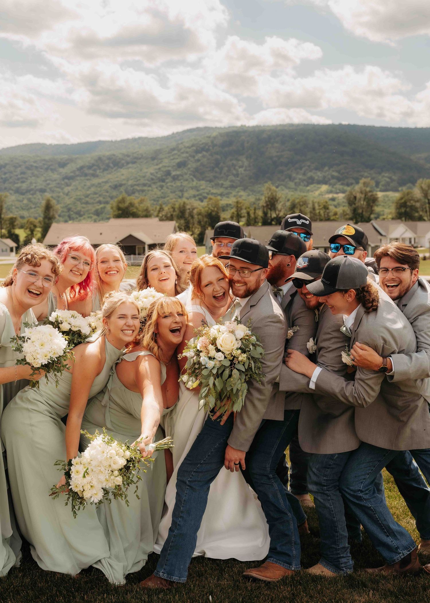 Joyful wedding party group photo with bride, groom, bridesmaids in sage green, and groomsmen in gray suits outdoors.