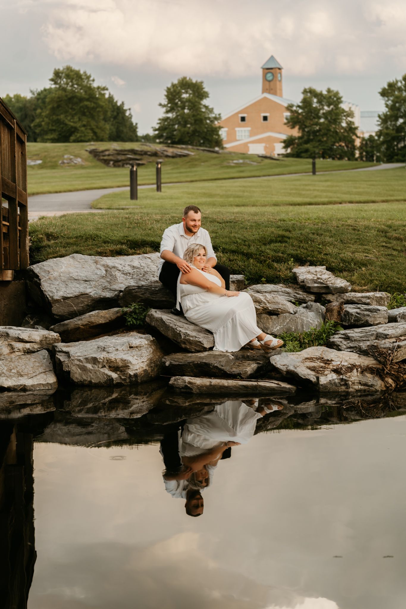 Couple sitting on rocks beside a reflective pond near a scenic park building at dusk.