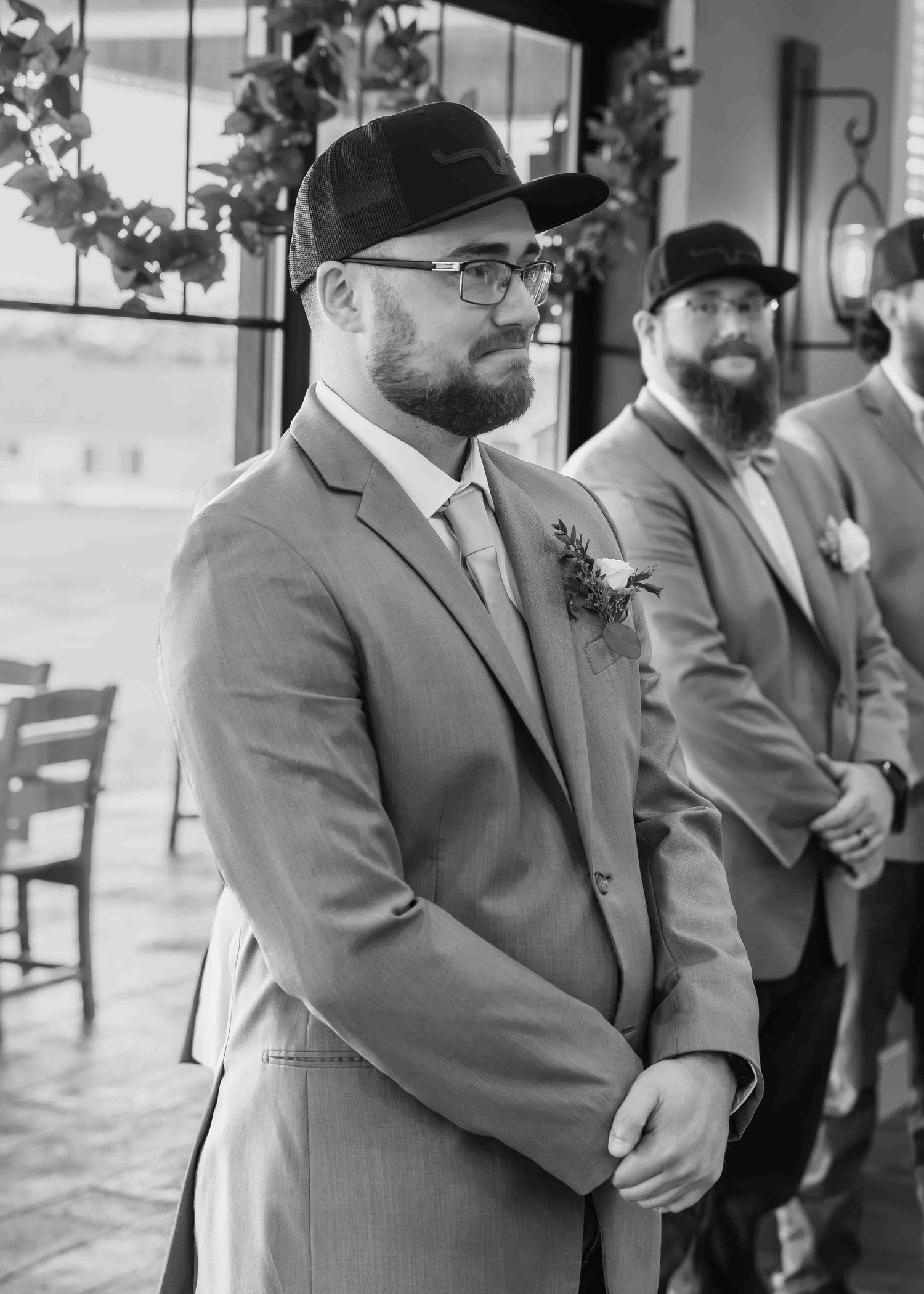 Groom in gray suit and black cap smiles at altar during wedding ceremony with groomsmen standing behind him.