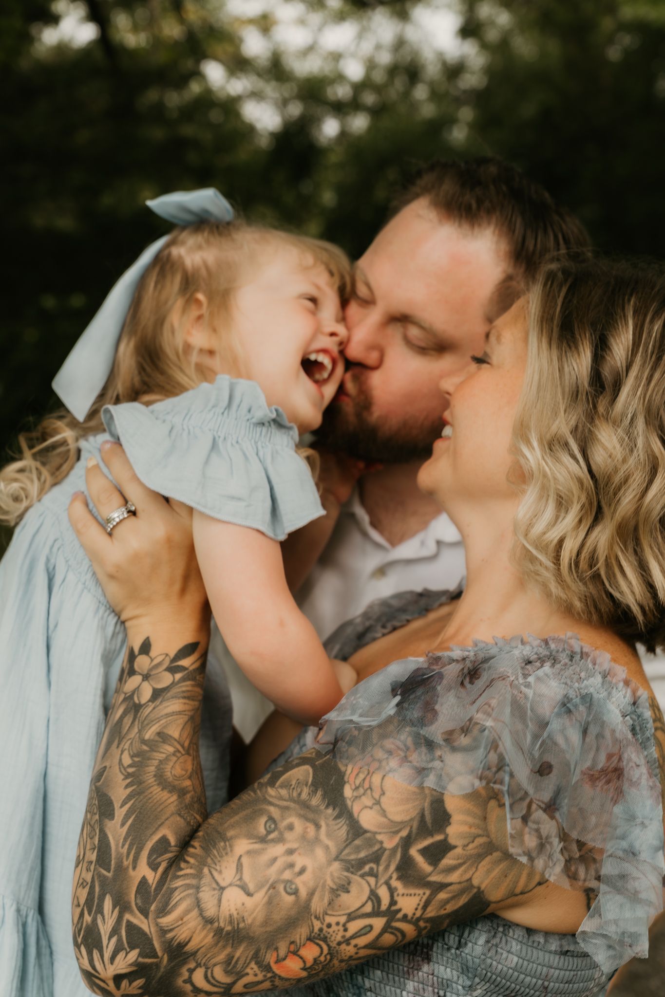 Tattooed parents kissing laughing toddler girl in blue bow dress outdoors in soft light.