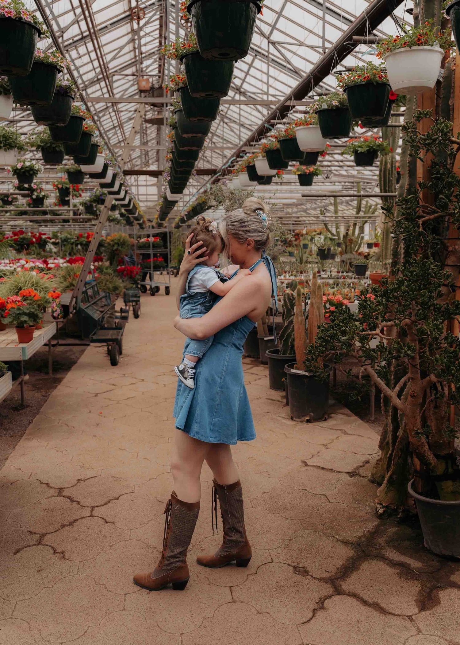 Mother in denim dress and cowboy boots hugging child in a lush greenhouse full of plants.