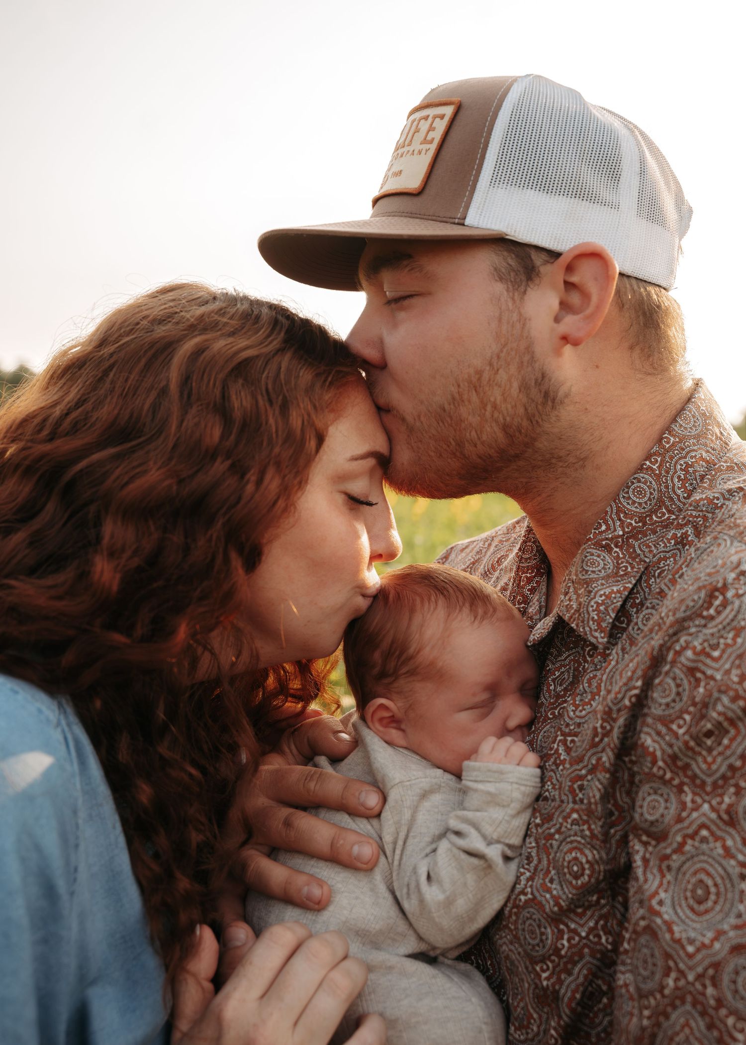 Young parents kissing their newborn baby outdoors in golden hour light, man wearing trucker hat and patterned shirt.