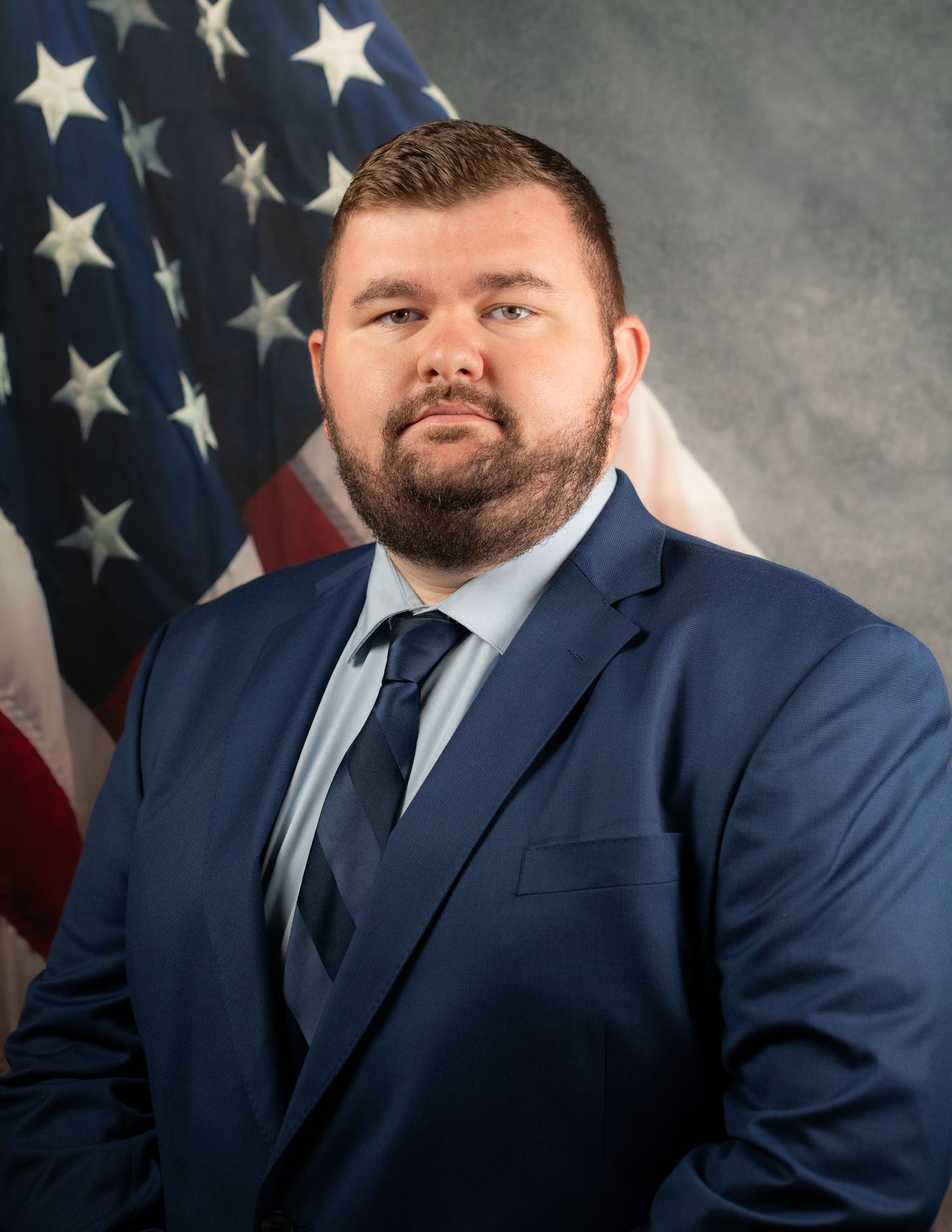Professional man in navy suit poses for portrait in front of American flag background.
