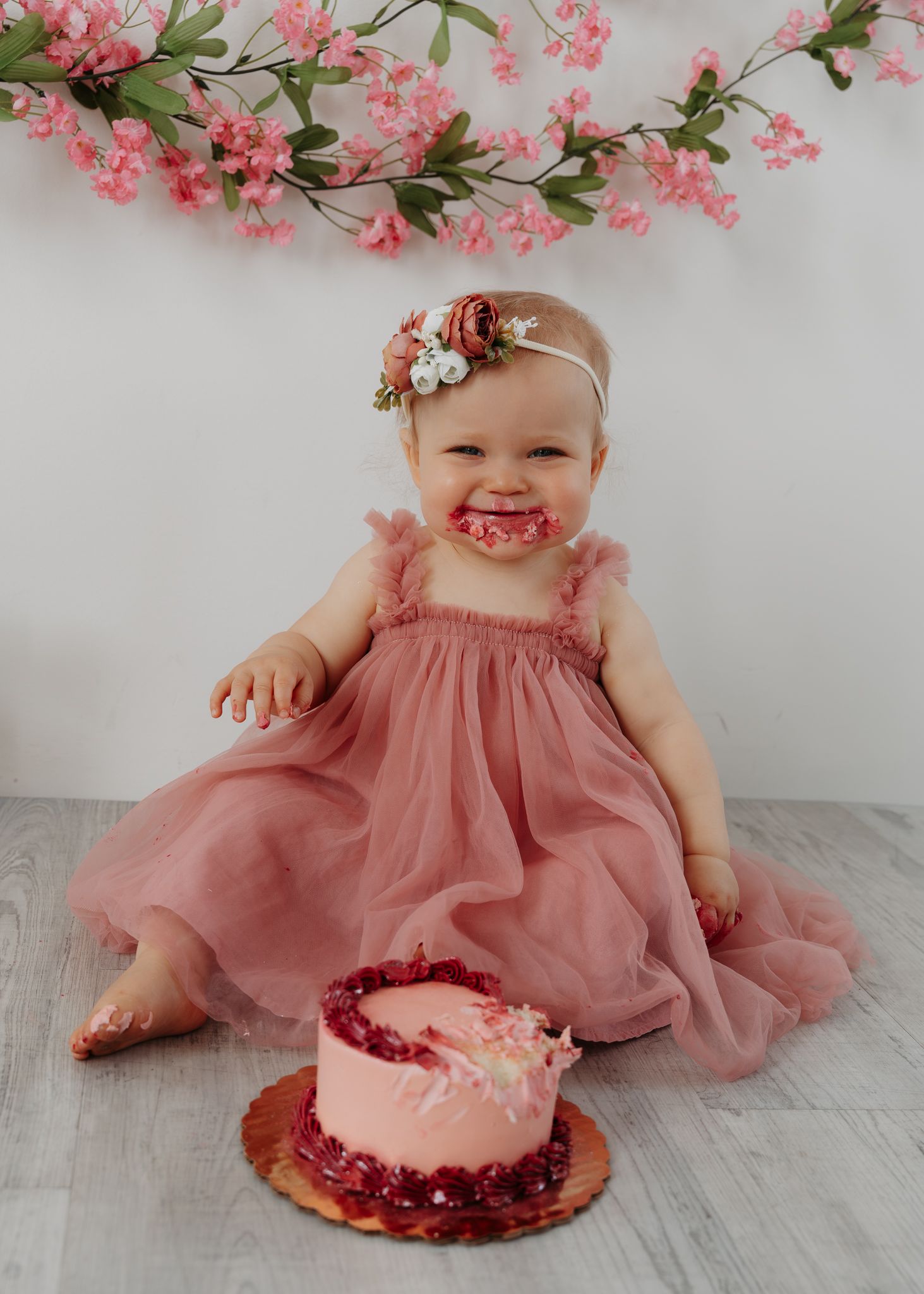 Smiling baby girl in a dusty rose tulle dress celebrates her first birthday smash cake session with floral backdrop.