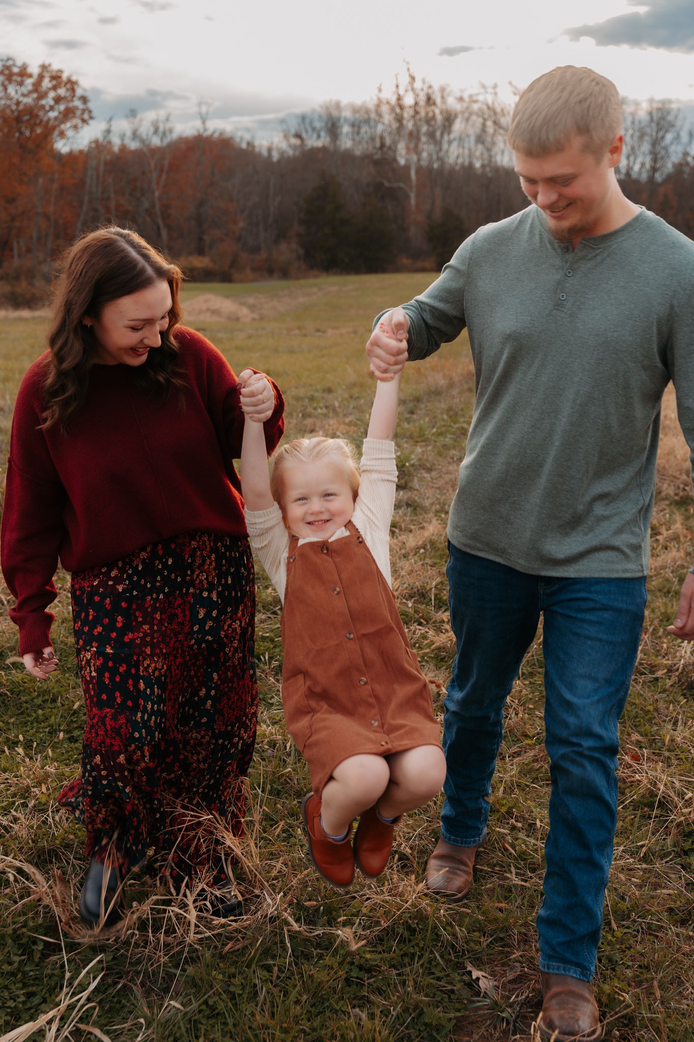 Parents swing their laughing toddler by the hands in an autumn field at golden hour.