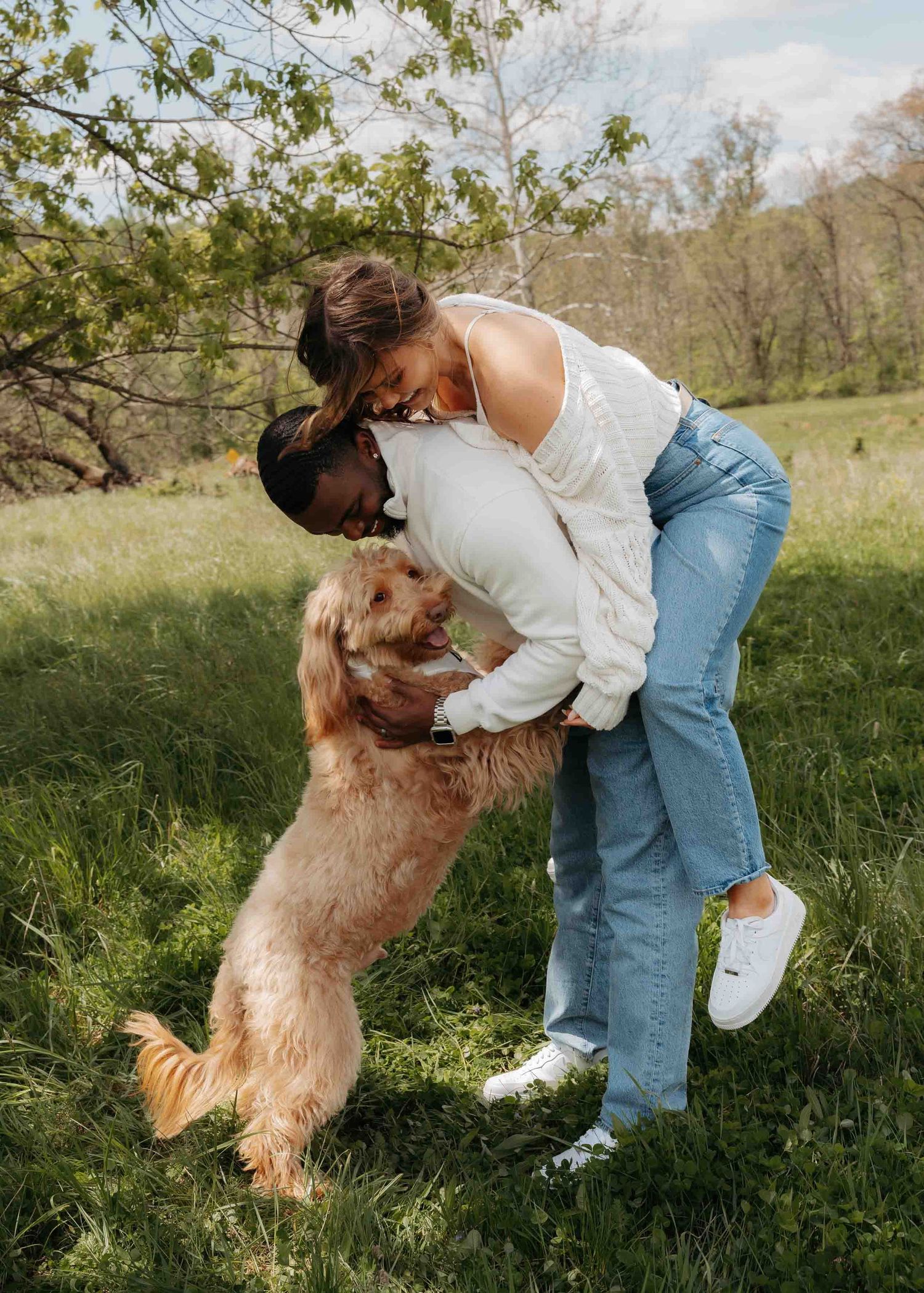 Couple sharing a tender moment while their golden doodle jumps up in a green park.
