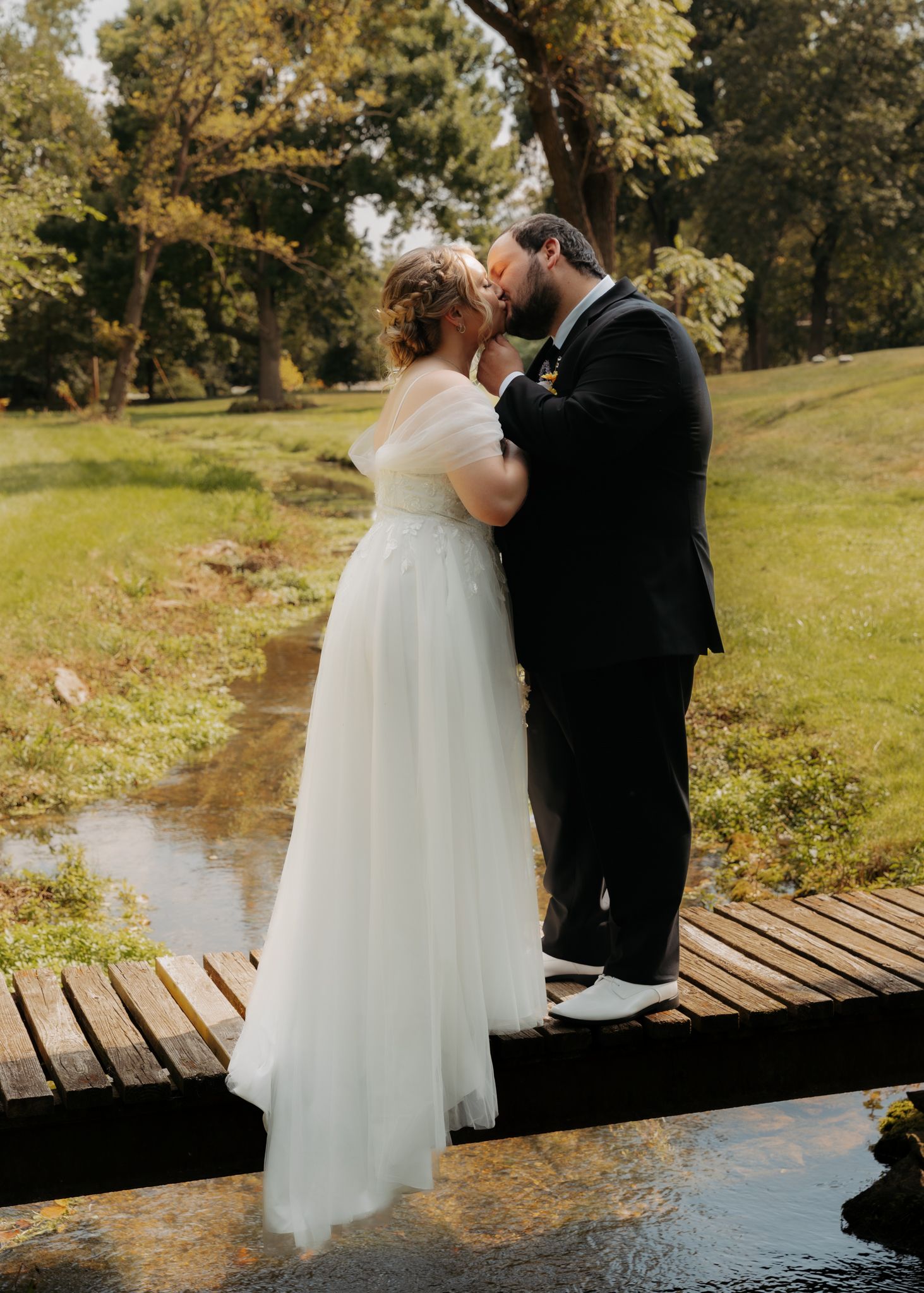 Newlyweds sharing a kiss on a wooden bridge over a stream in a lush green park.