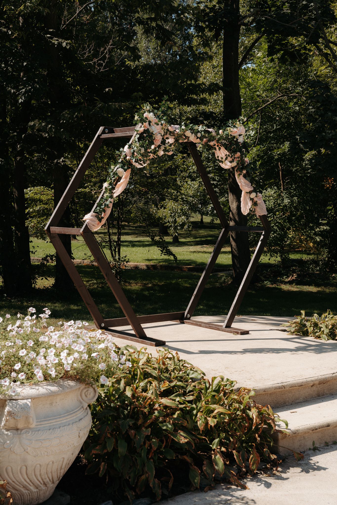 Geometric hexagonal wooden wedding arch decorated with blush flowers in an outdoor garden setting.