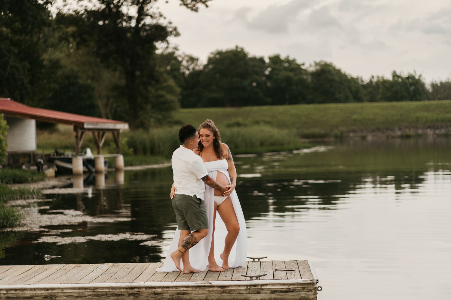 A couple shares a romantic moment on a wooden dock by a serene lake surrounded by lush green trees.