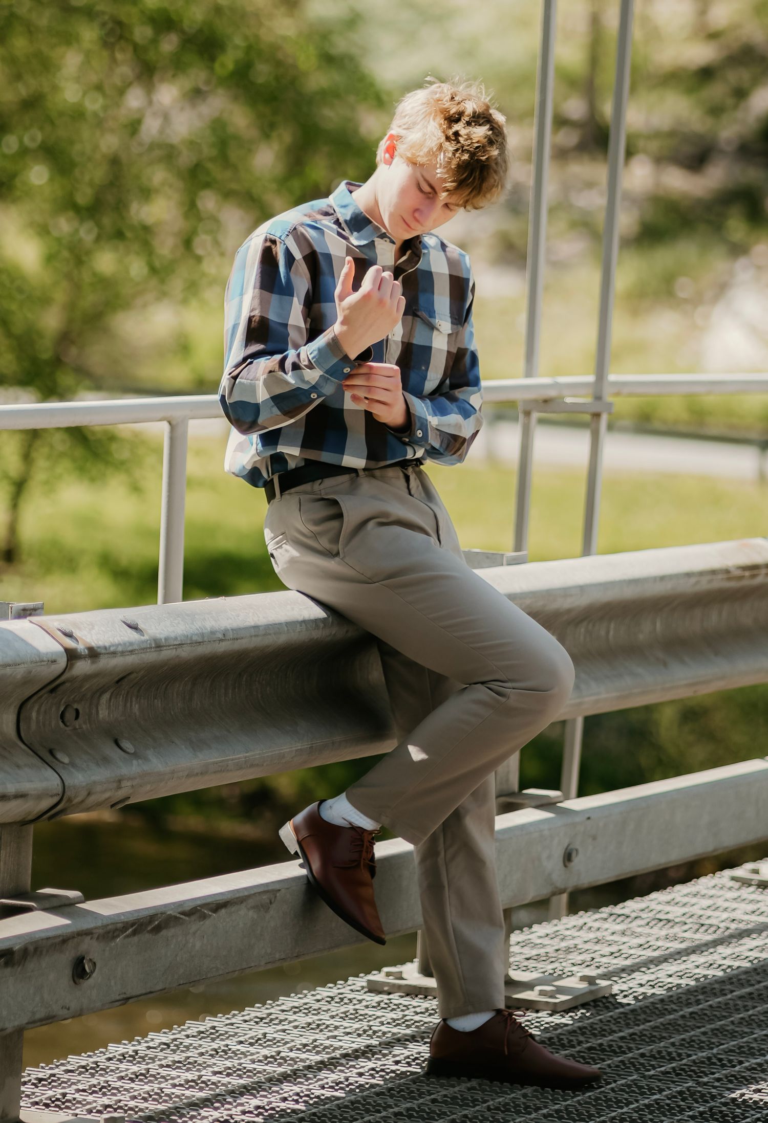 Young man in plaid shirt and khaki pants leans on metal railing in sunny outdoor setting.