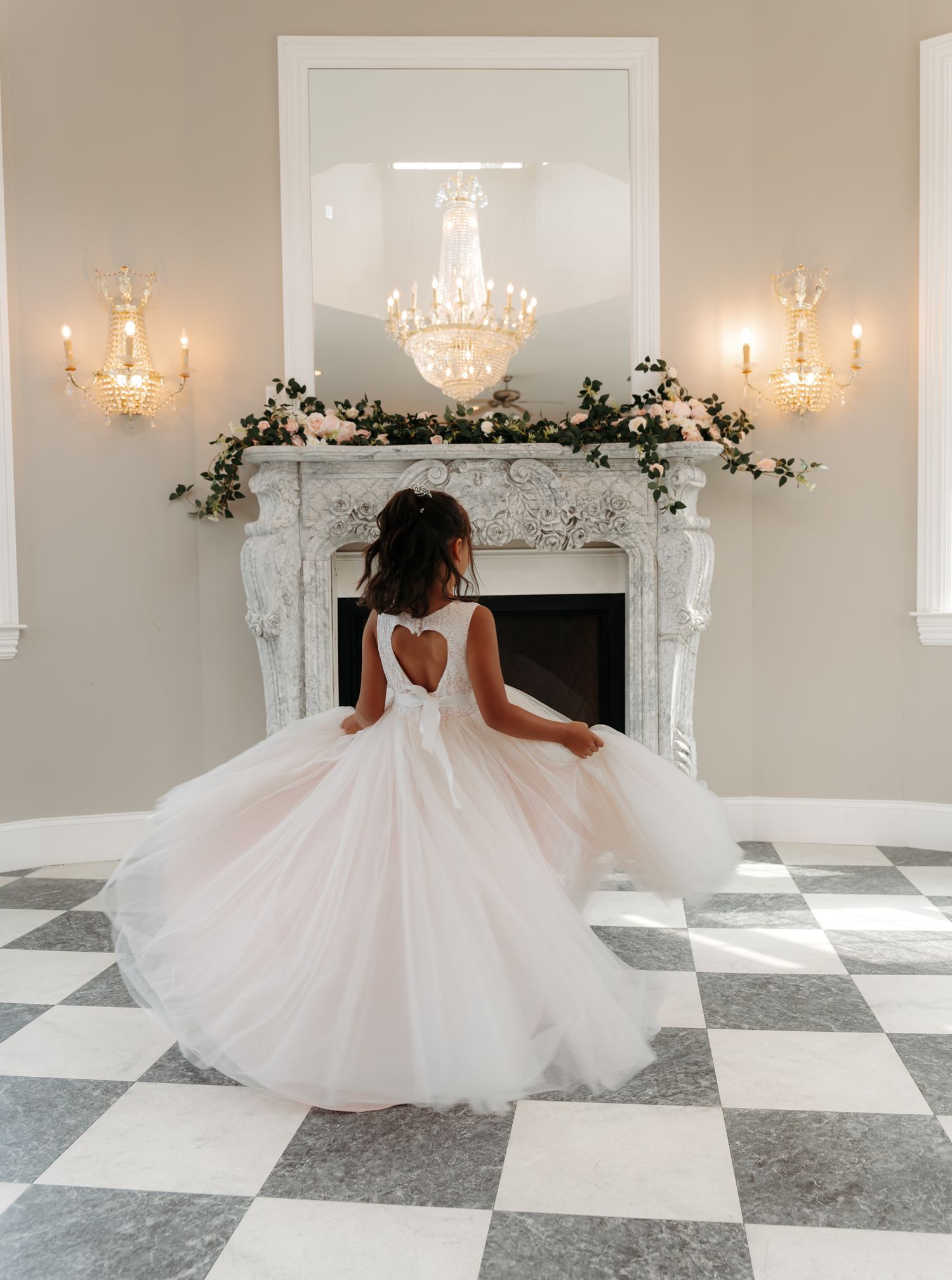 Flower girl in blush tulle dress twirls before elegant fireplace with chandelier in wedding venue ballroom.