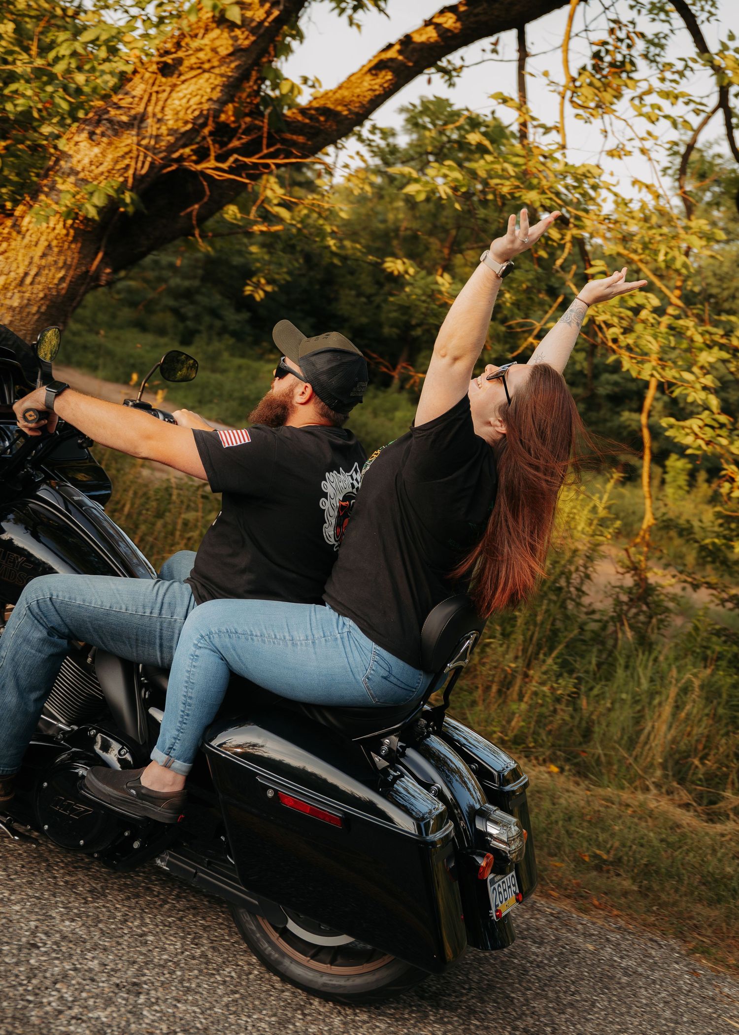 Two people on a Harley-Davidson motorcycle raise their arms joyfully under golden sunlight trees on a scenic road.