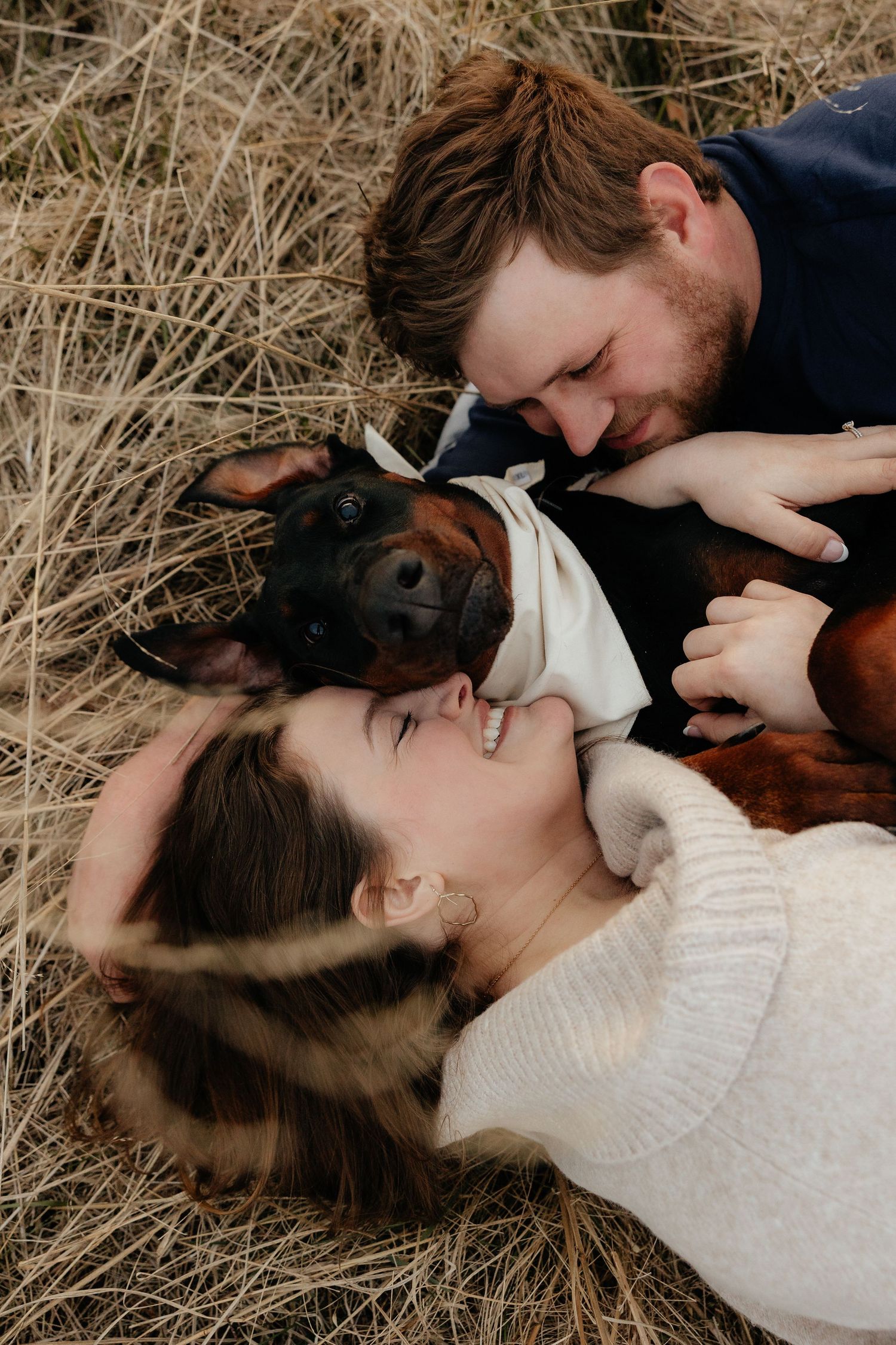 Couple lying in dry hay field smiling at each other with their Doberman dog between them.