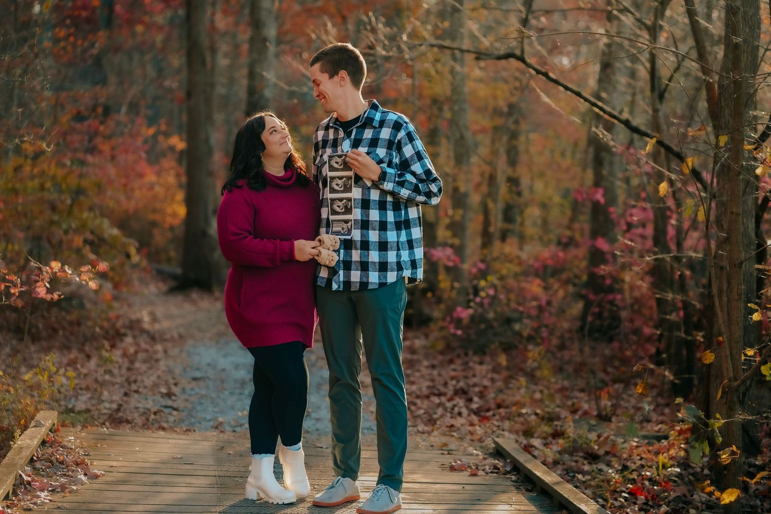 Couple holding ultrasound photos on a woodland boardwalk during fall pregnancy announcement.