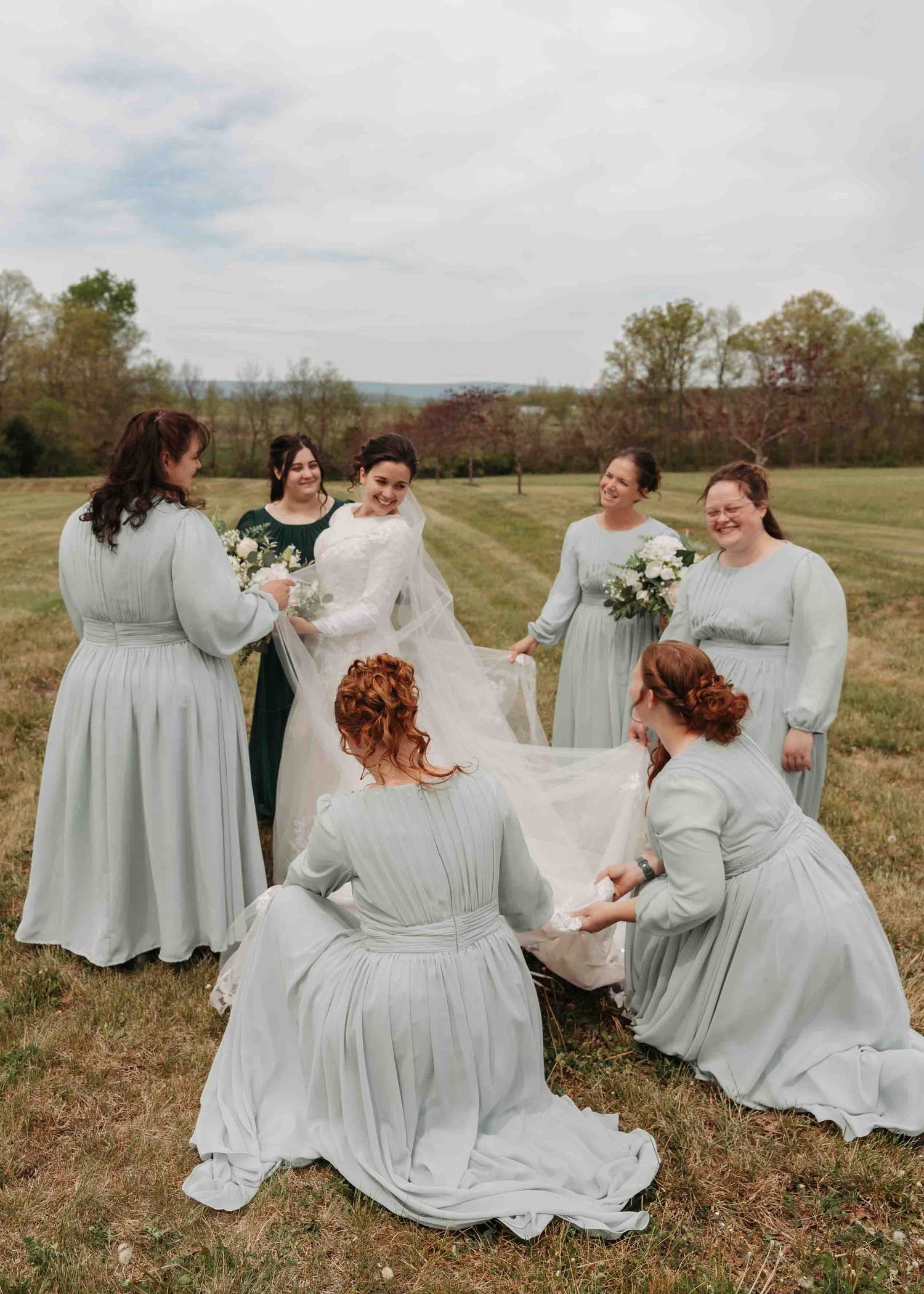 Bride surrounded by bridesmaids in sage green dresses holding bouquets in an outdoor field.