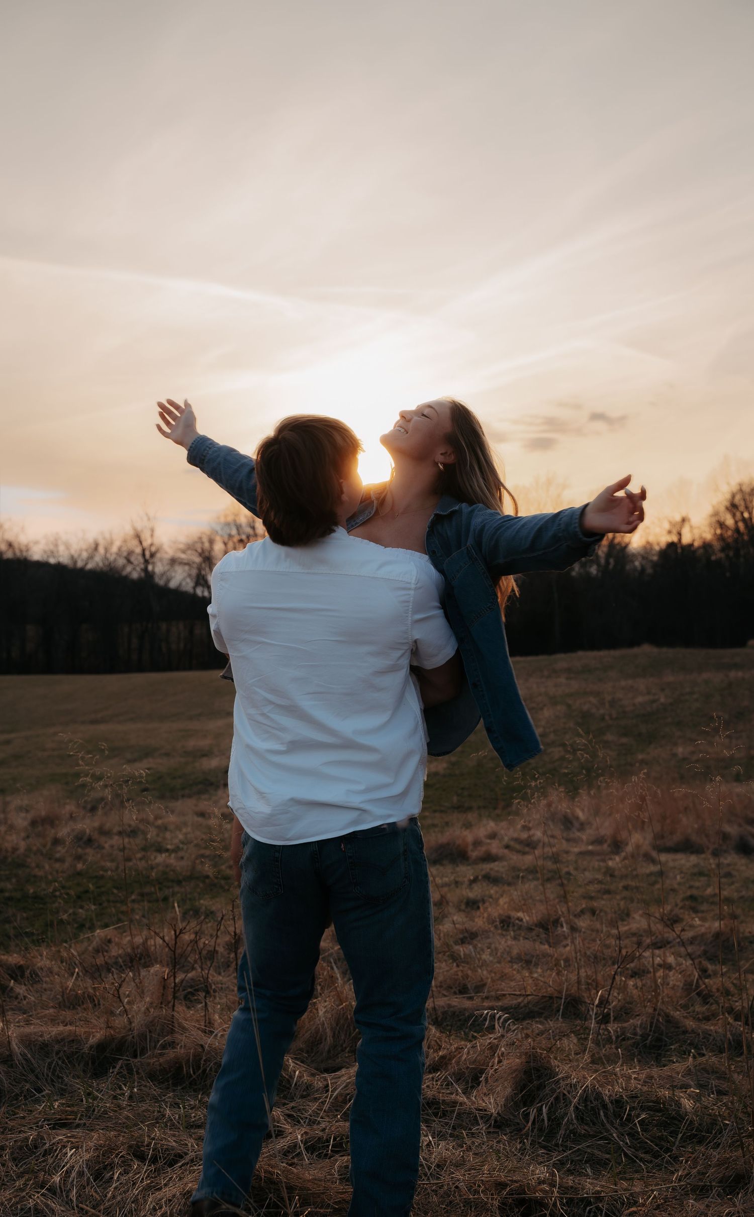 Man lifting woman with outstretched arms in an open field during a glowing sunset.