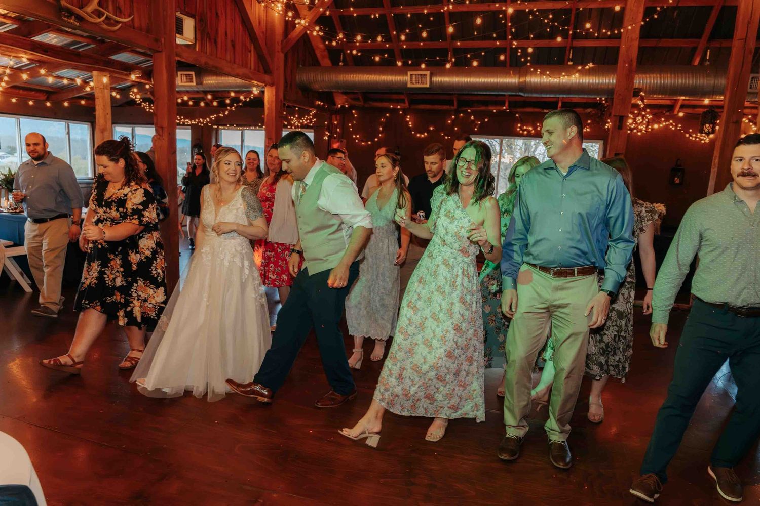 Wedding guests line dancing in a rustic barn venue decorated with string lights during a lively reception.