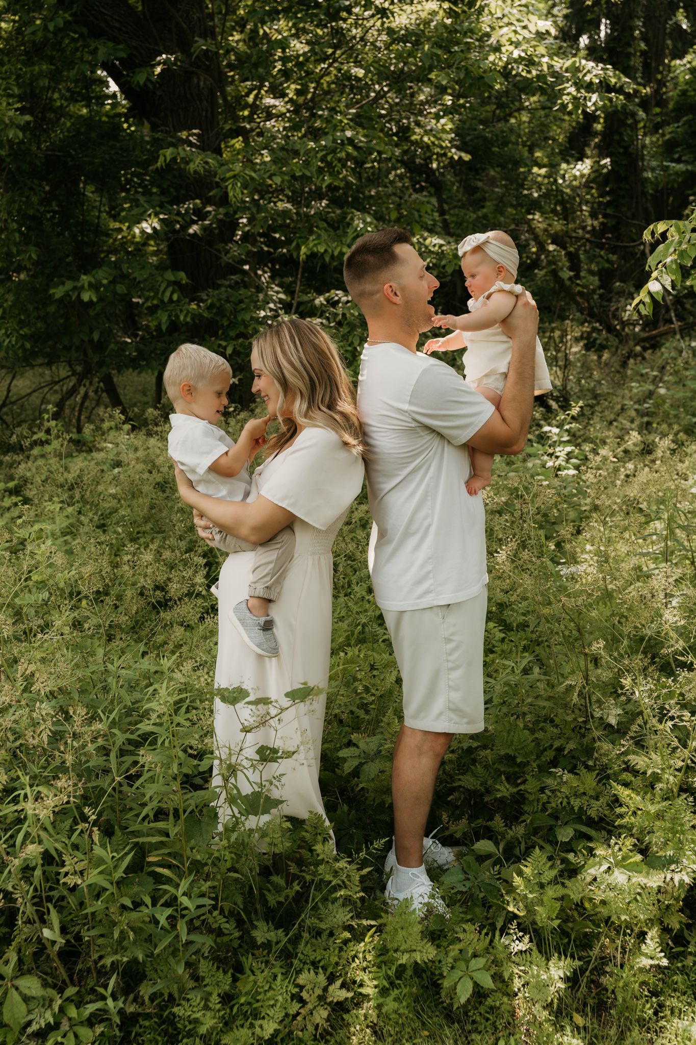 Young couple in matching white outfits holding two babies in a lush green forest setting.