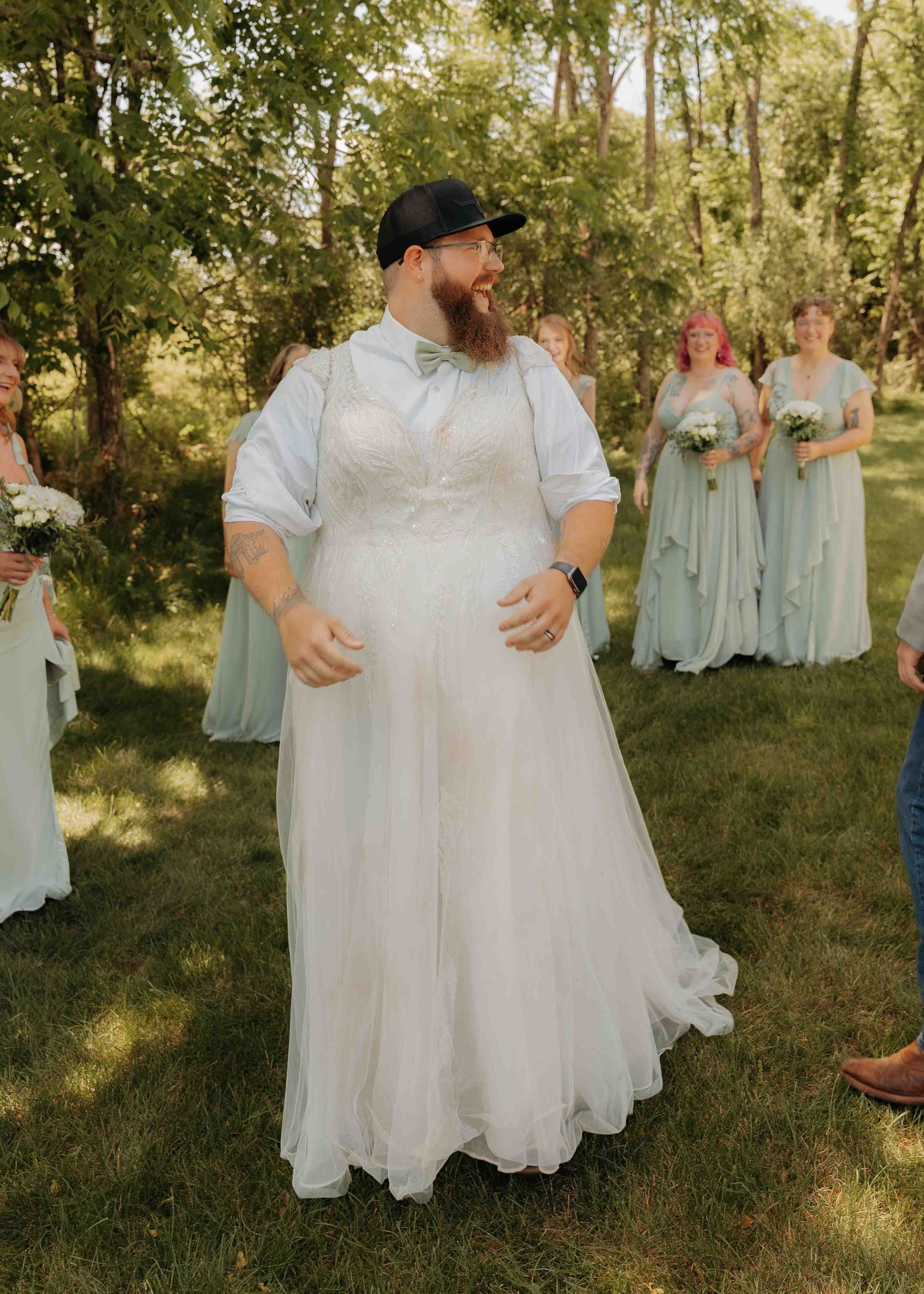 Bearded groom in white wedding dress and bow tie stands smiling with bridesmaids in sage green gowns outdoors.