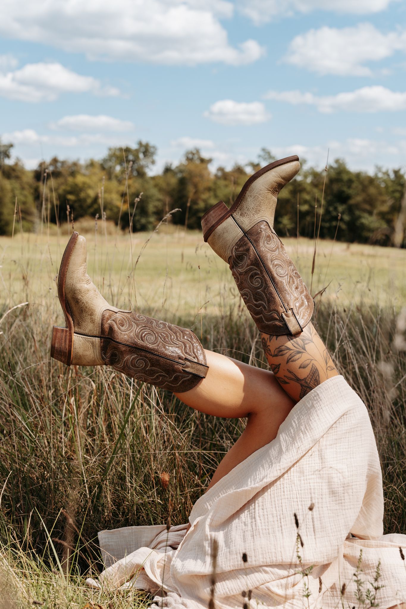 Person sitting in a field wearing decorative cowboy boots with tattoos, legs raised against a blue sky.