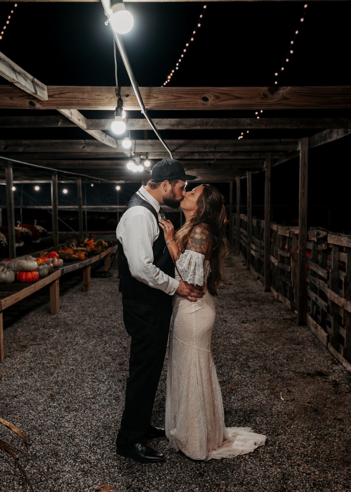 A couple shares a romantic kiss under string lights in a rustic wooden structure decorated with pumpkins and fall decor.