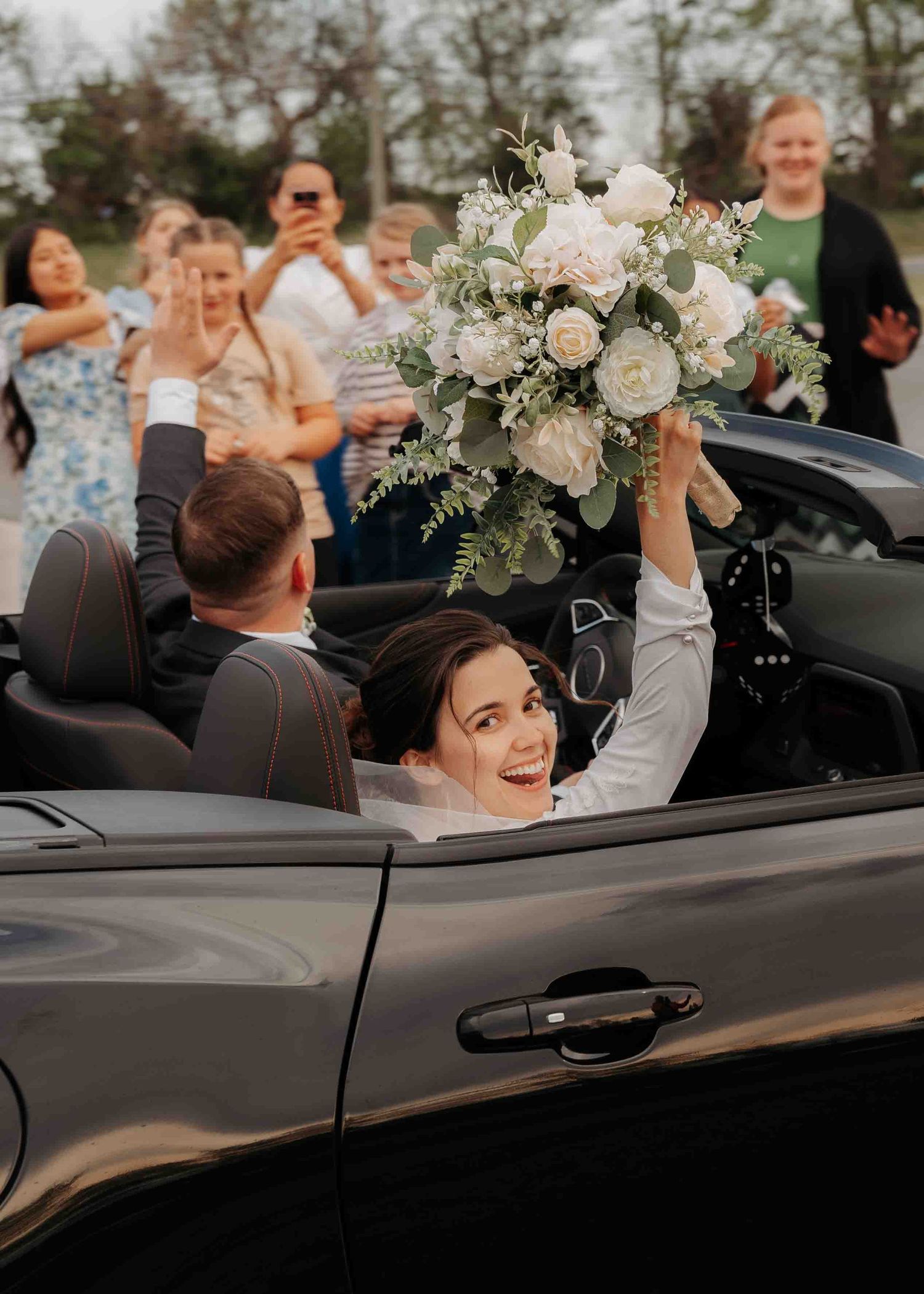Joyful bride raises white floral bouquet while departing in a convertible car.